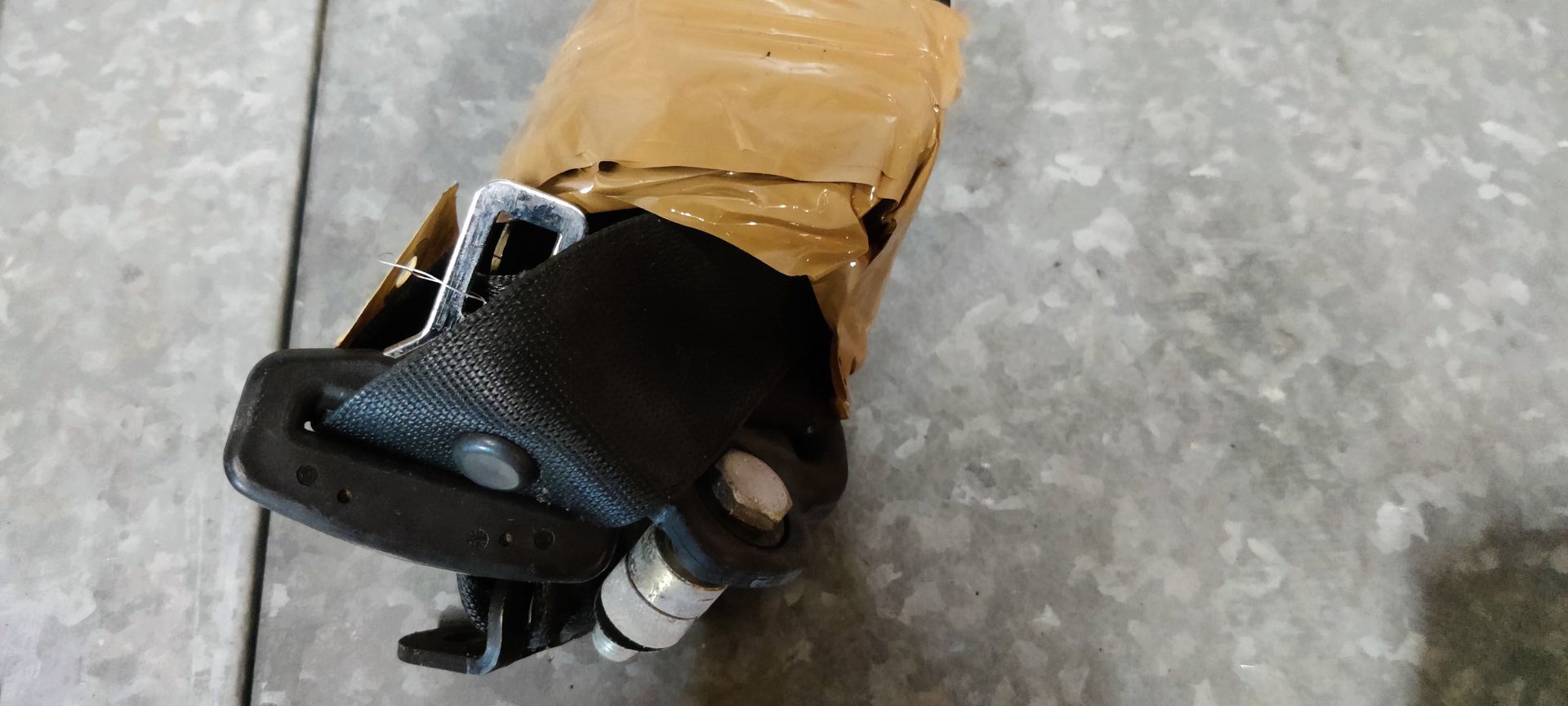 Een zwarte veiligheidsgordel, gedeeltelijk omwikkeld met bruine verpakkingstape, ligt op een grijs metalen oppervlak.