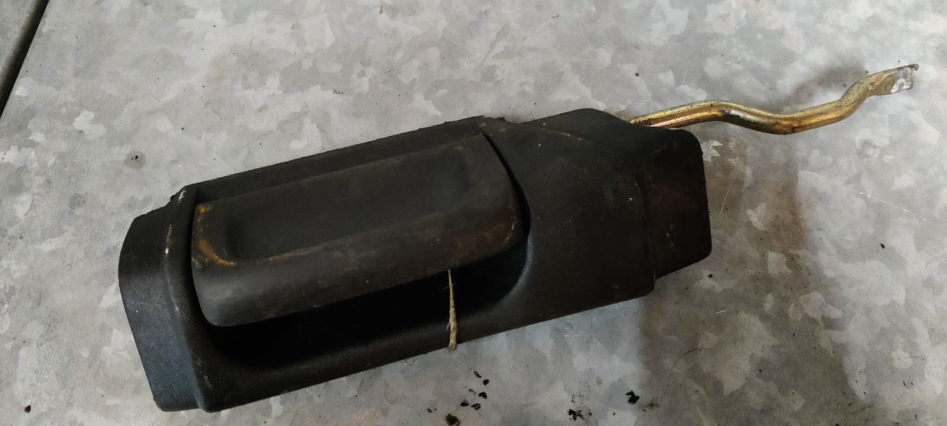 Een zwarte autodeurhendel met een metalen verbindingsstang, liggend op een grijze ondergrond.