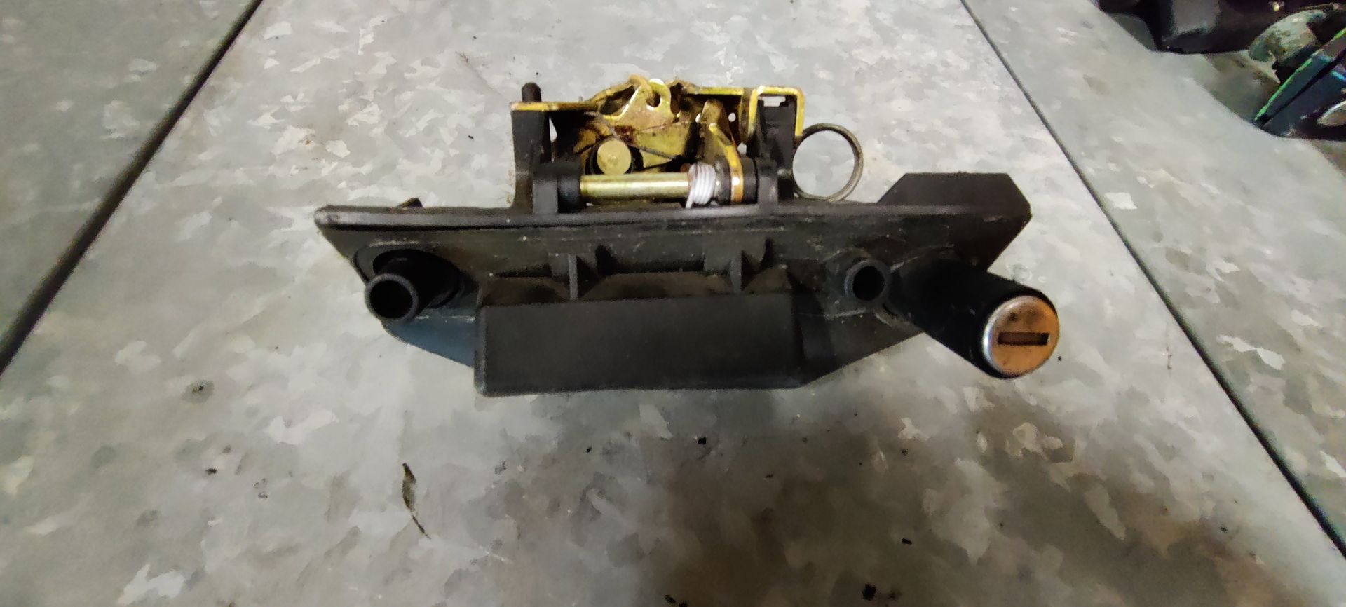 Een zwart autoslot met ingebouwde cilinder voor de kofferbak of achterklep, gezien vanaf de achterkant op een metalen bureau.