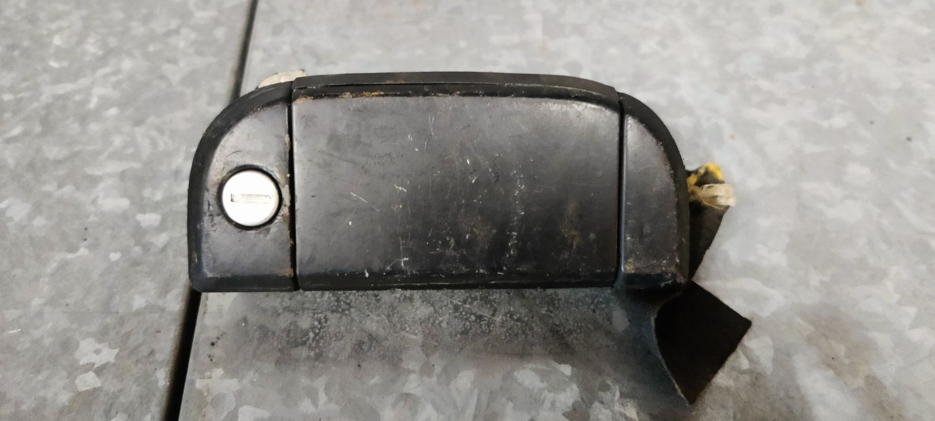 Een zwarte, rechthoekige autodeurhendel met een sleutelgat aan de linkerkant, afgebeeld tegen een grijze metalen achtergrond.
