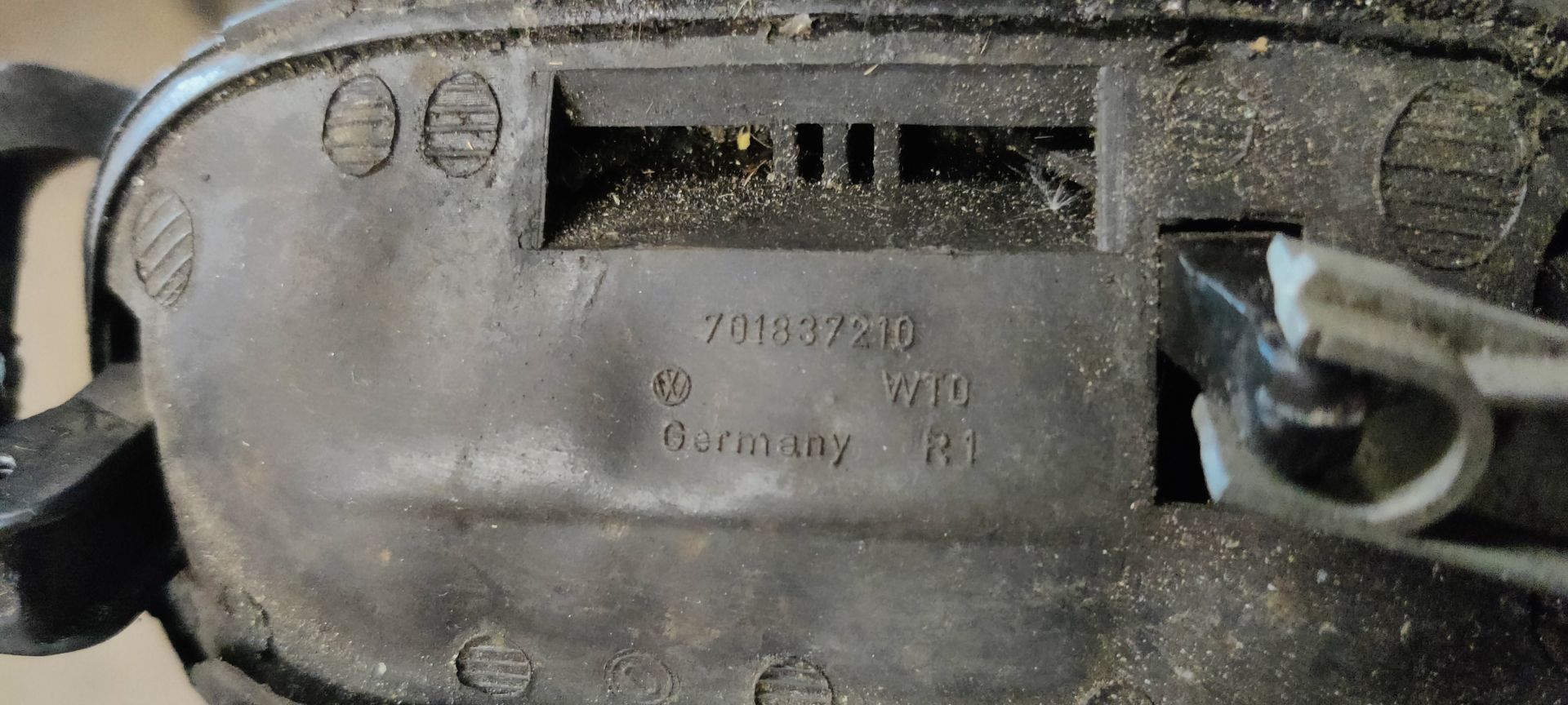 Een zwarte, rechthoekige autodeurhendel met een sleutelgat aan de linkerkant, afgebeeld tegen een grijze metalen achtergrond.