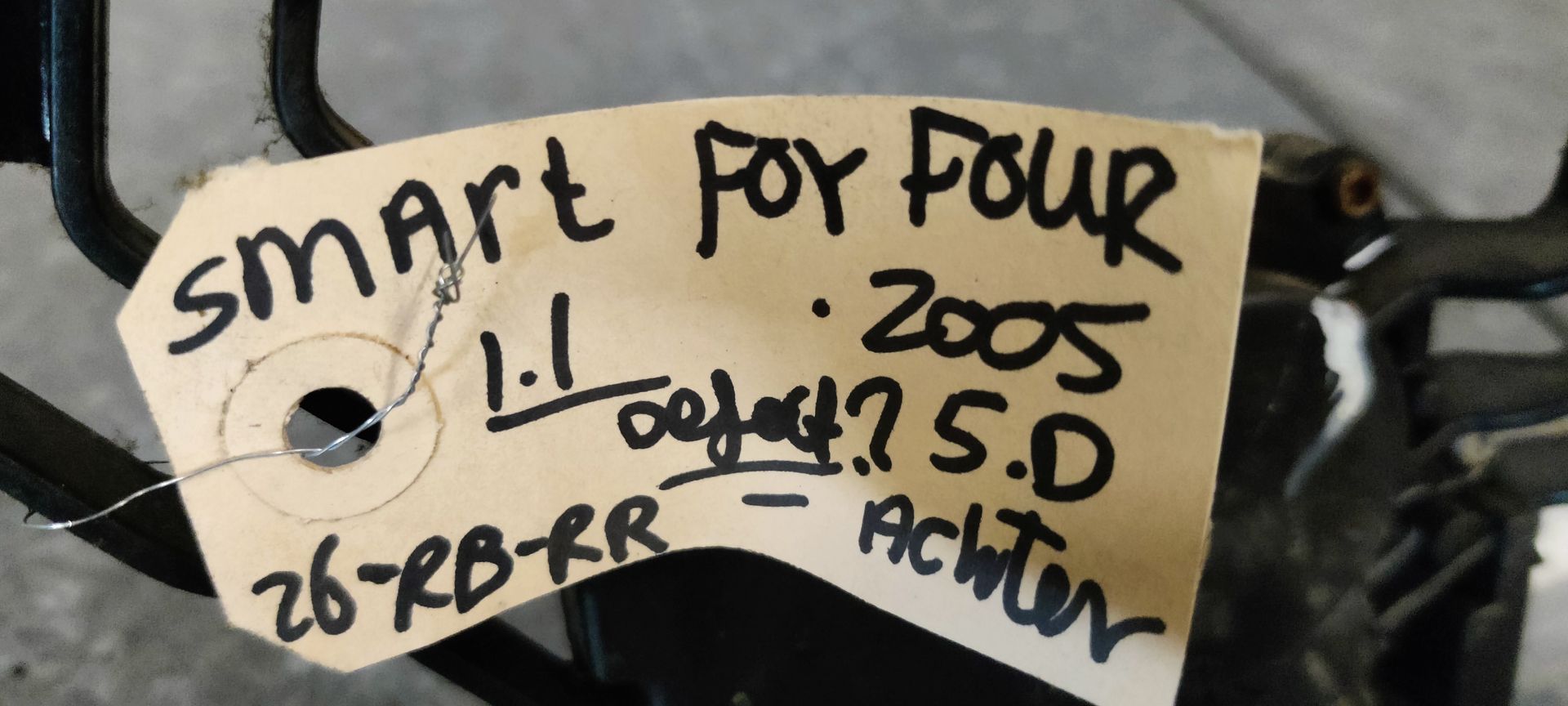 Een zilverkleurige ruitenwissermotor met een zwarte montagebeugel en identificatielabel op een grijze ondergrond.