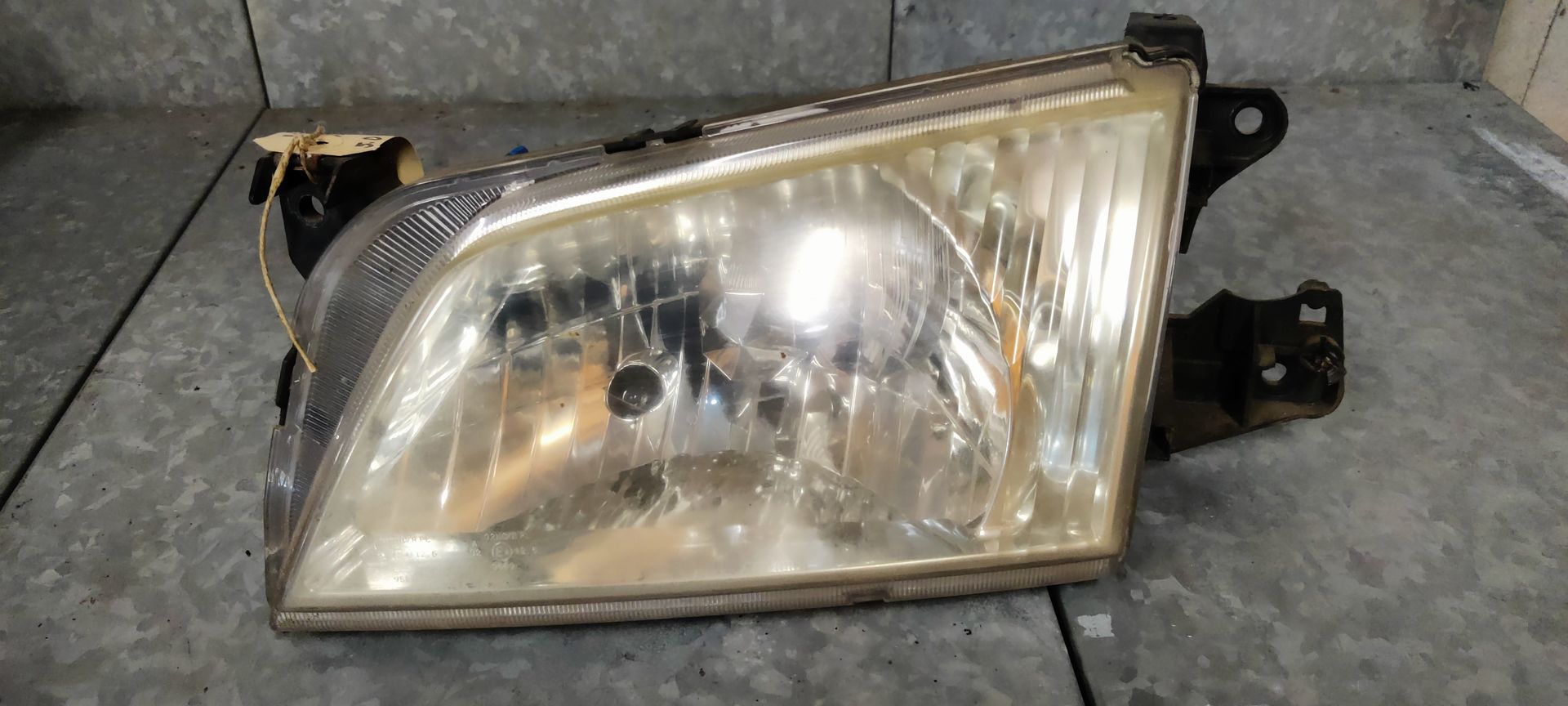 Een vergeelde, gebruikte rechthoekige autokoplampunit met zwarte plastic bevestigingsbeugels op een grijze ondergrond.