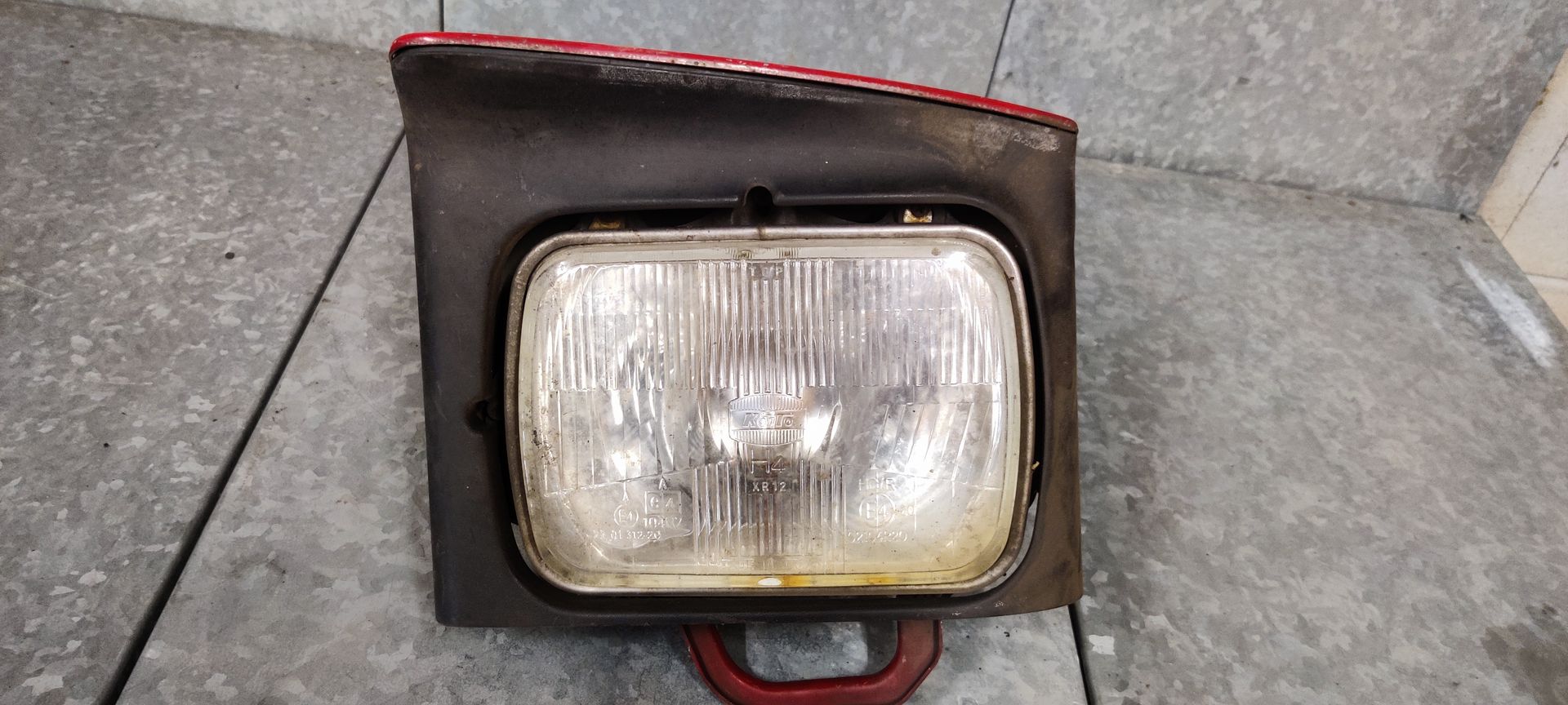 Een rechthoekige, verweerde autokoplampunit met een verchroomde glazen lens in een donkergrijze kunststof behuizing.