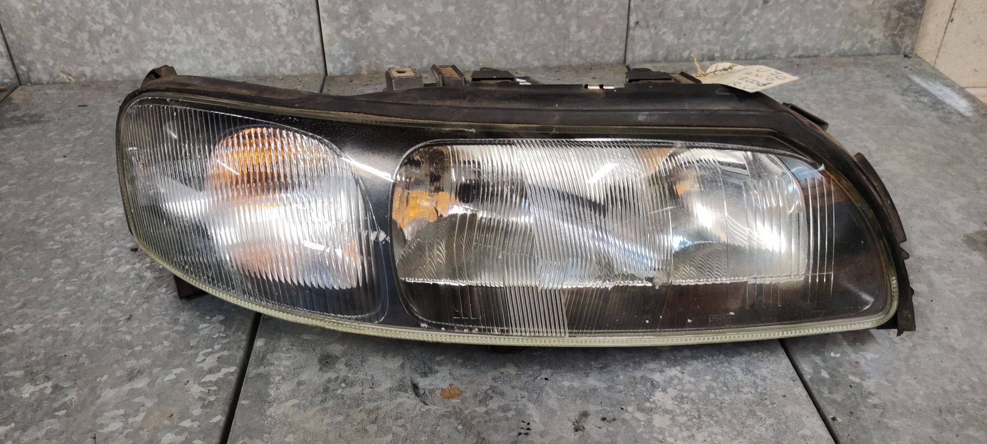 Een vervangende autokoplampunit met een heldere lens en een zwarte behuizing, geplaatst tegen een grijze tegelachtergrond.