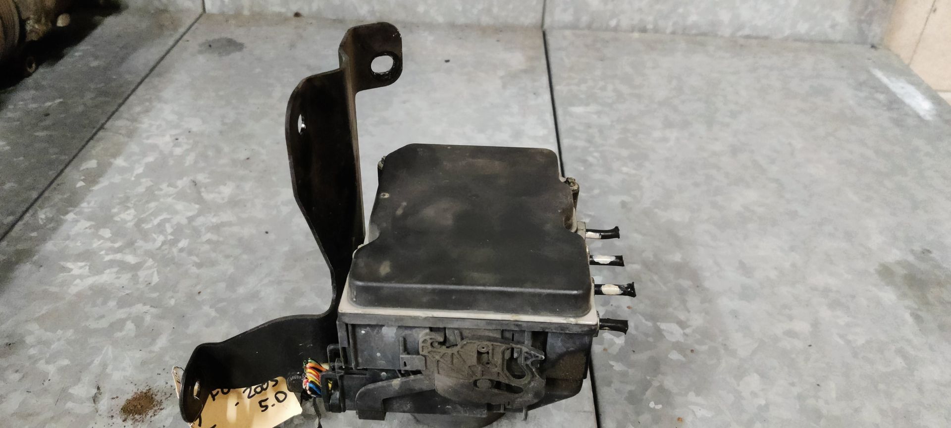 Een metalen motorregelunit voor auto's met een zwarte montagebeugel op een grijze betonnen ondergrond.