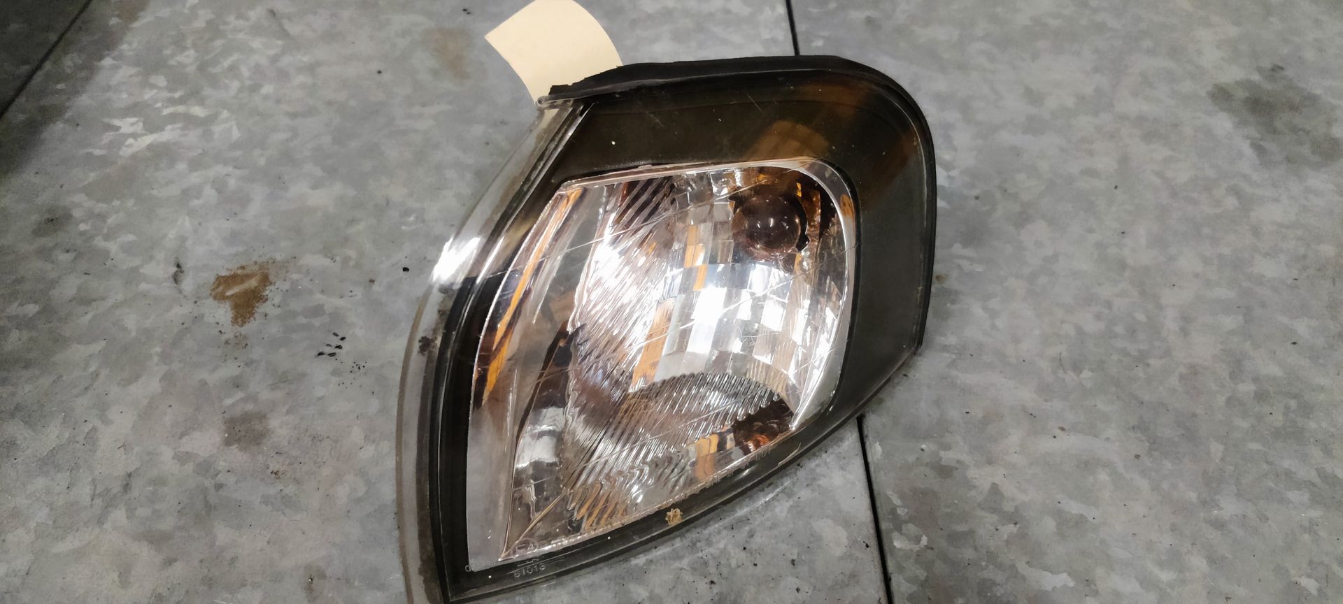 Een enkele autokoplampunit met een zwarte rand en een transparante reflectorlens, rustend op een grijze betonnen vloer.