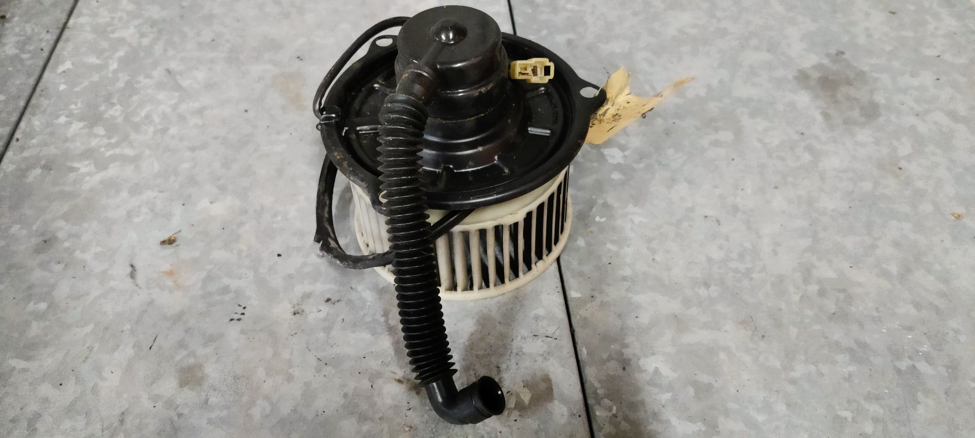 Een zwarte autoventilator met een wit ventilatorwiel en een flexibele zwarte koelslang staat op een grijze betonnen vloer.