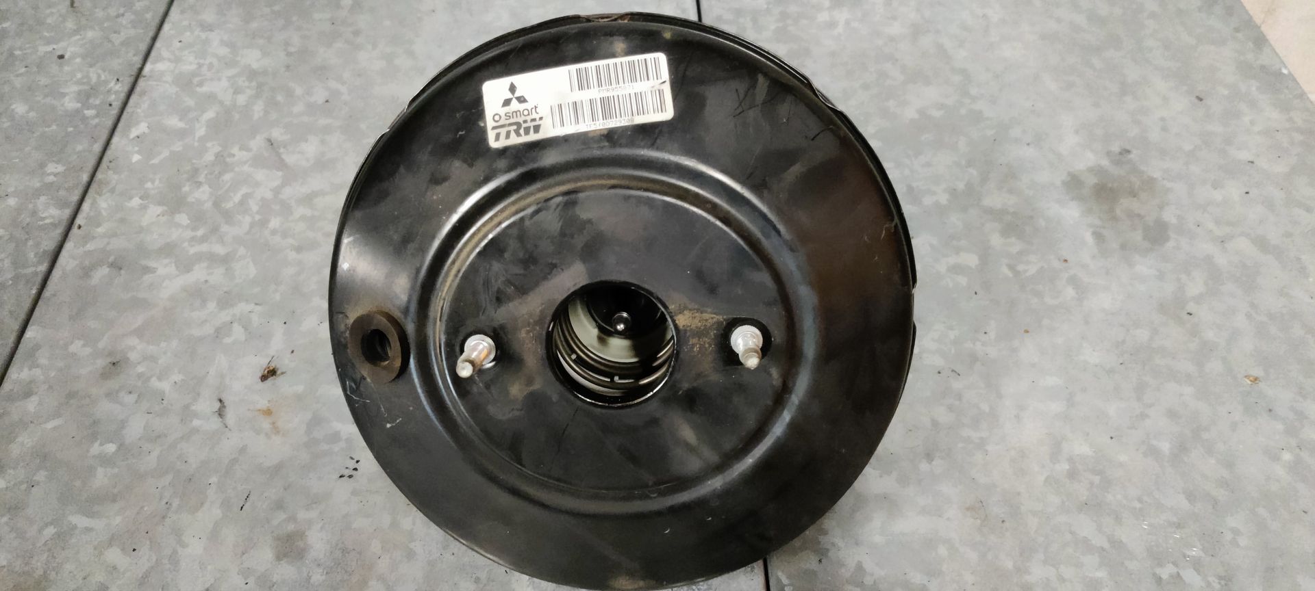 Een zwarte Mitsubishi-rembekrachtiger met een wit identificatielabel, van achteren gezien op een metalen oppervlak.