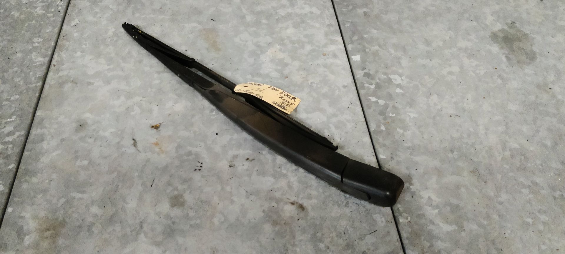 Een zwarte ruitenwisserarm met een aangehecht wisserblad ligt op een grijze betonnen ondergrond.