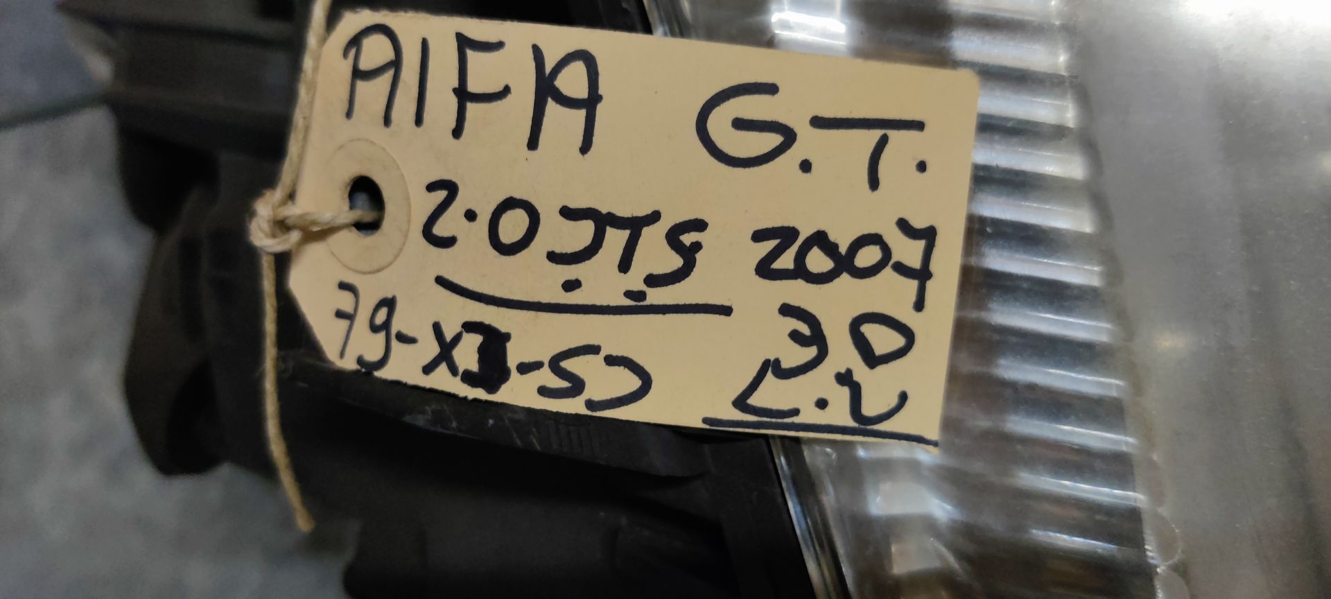 Een gebruikte koplampunit met een handgeschreven papieren label eraan, liggend op een grijze ondergrond.