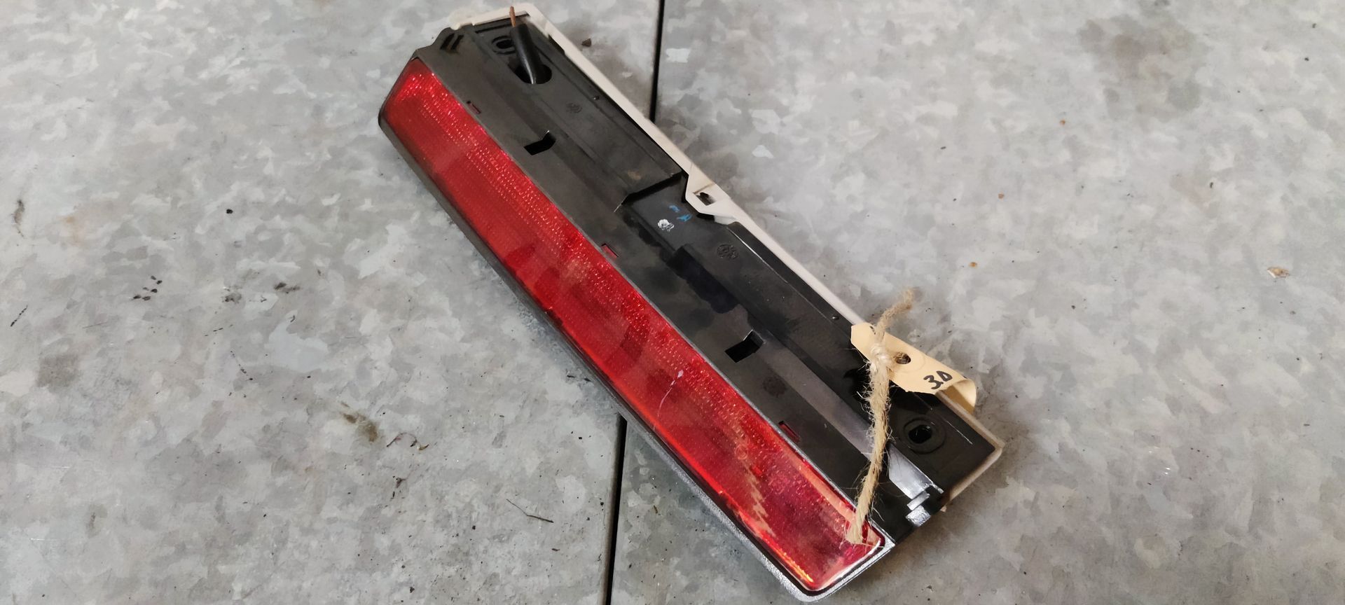 Een rood-zwarte achterlichtunit van een auto, met een klein identificatielabeltje eraan, ligt op een grijze betonnen vloer.