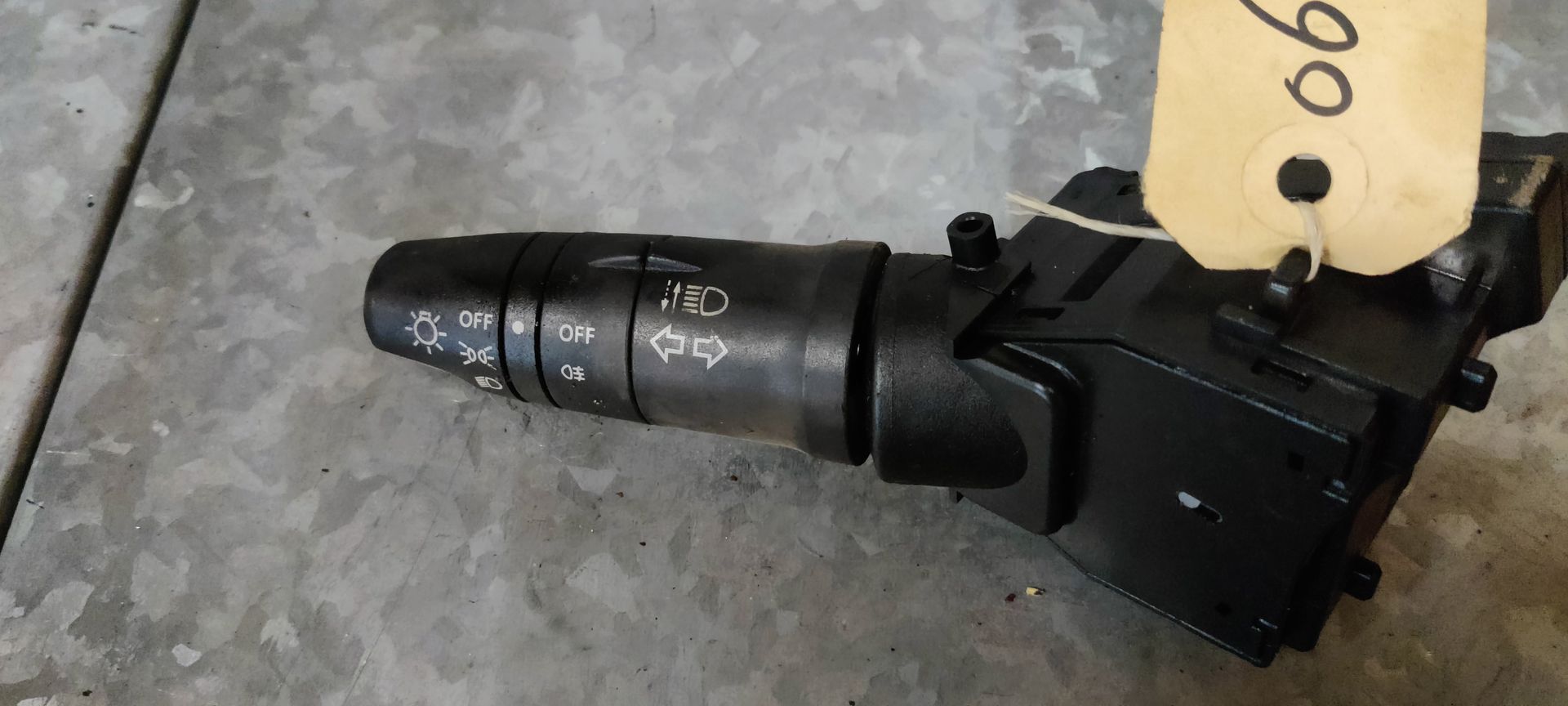 Een zwarte stuurkolomhendel voor richtingaanwijzers en verlichting met witte pictogrammen, rustend op een metalen oppervlak.
