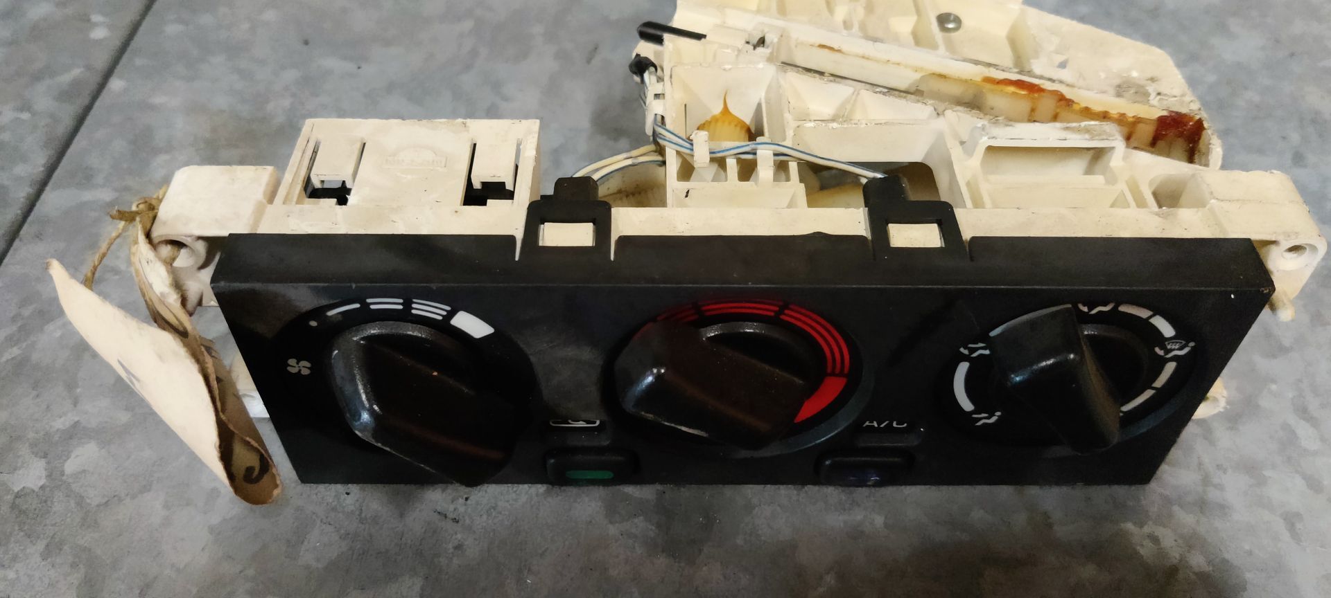 Een klimaatregelingsunit voor auto's met drie zwarte draaiknoppen en een witte kunststof behuizing op een grijze ondergrond.