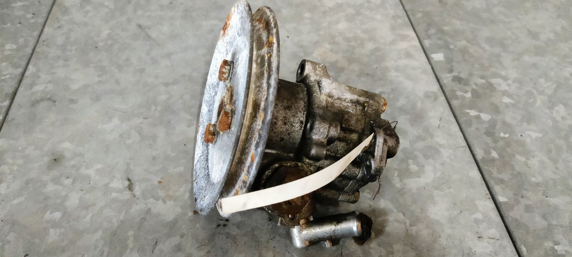 Een versleten, vuile waterpomp van een auto met een verroeste metalen poelie, staat op een tegelvloer.