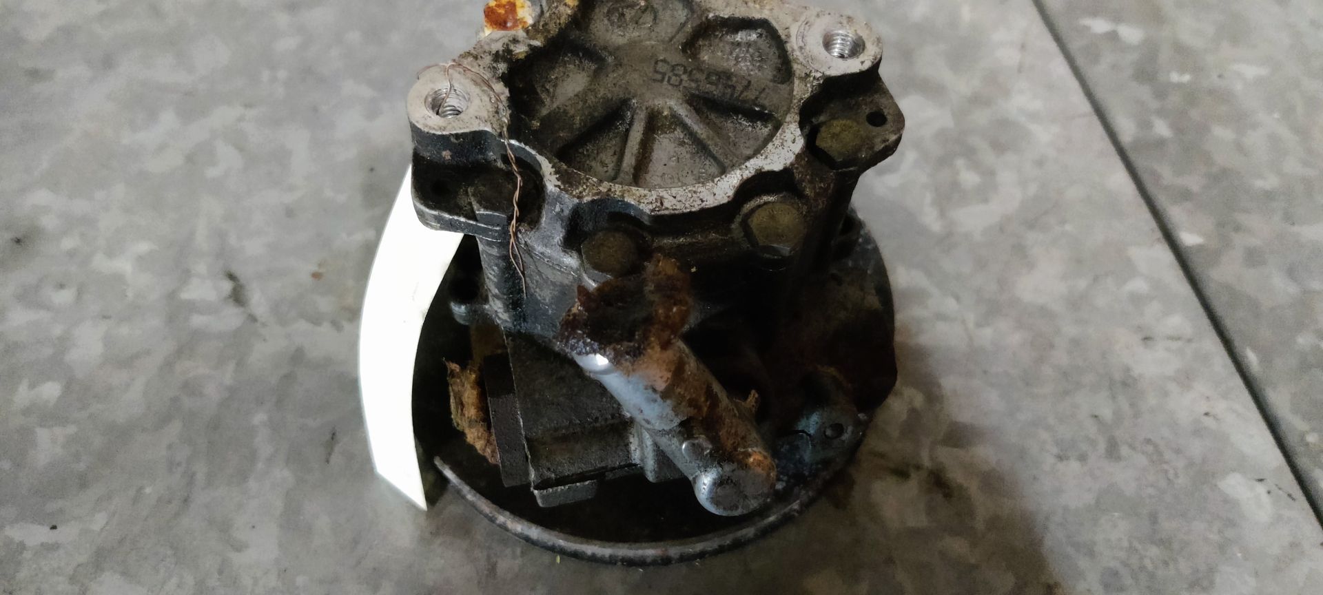 Een versleten, vuile waterpomp van een auto met een verroeste metalen poelie, staat op een tegelvloer.