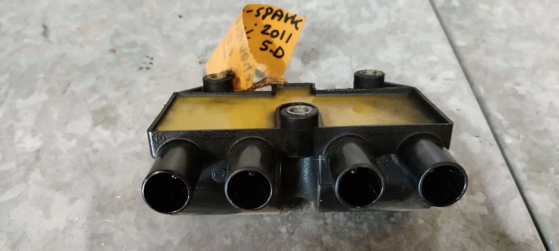 Een zwarte bobine voor een Chevrolet Spark uit 2011, met vier aansluitingen voor bougiekabels en een geel identificatielabel.
