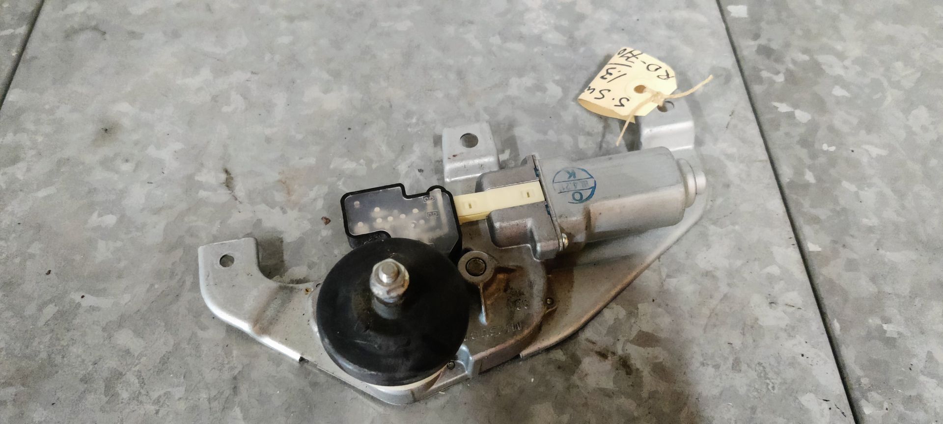Een metalen raammotor voor een auto, met een zwart plastic tandwiel, rustend op een grijze tegelondergrond.