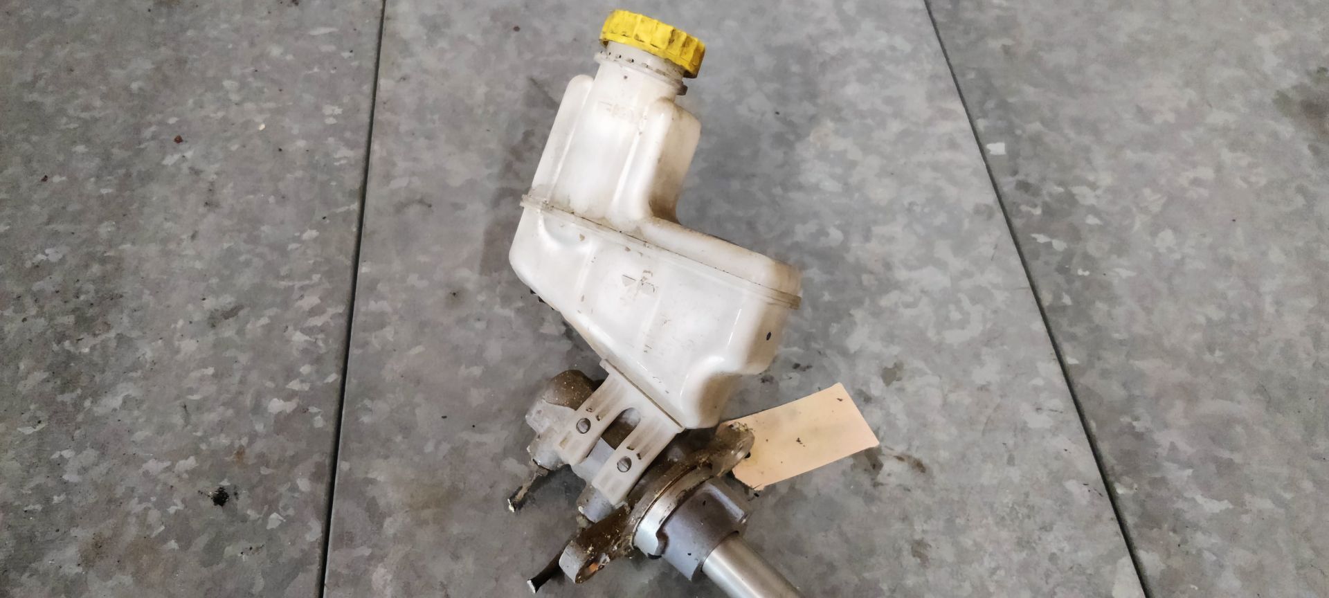 Een hoofdremcilinder van een auto met een doorschijnend reservoir en een gele dop, rustend op een grijze betonnen ondergrond.