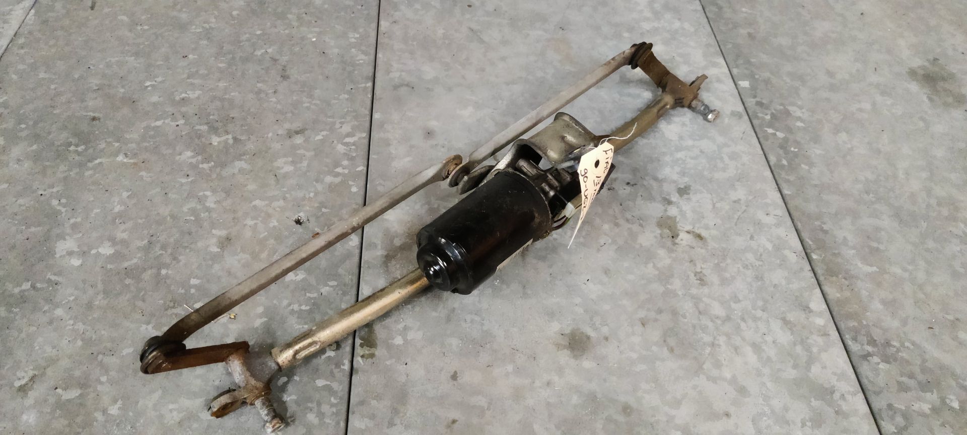 Een ruitenwissermotor en -mechanisme van een auto liggen op een betonnen ondergrond.