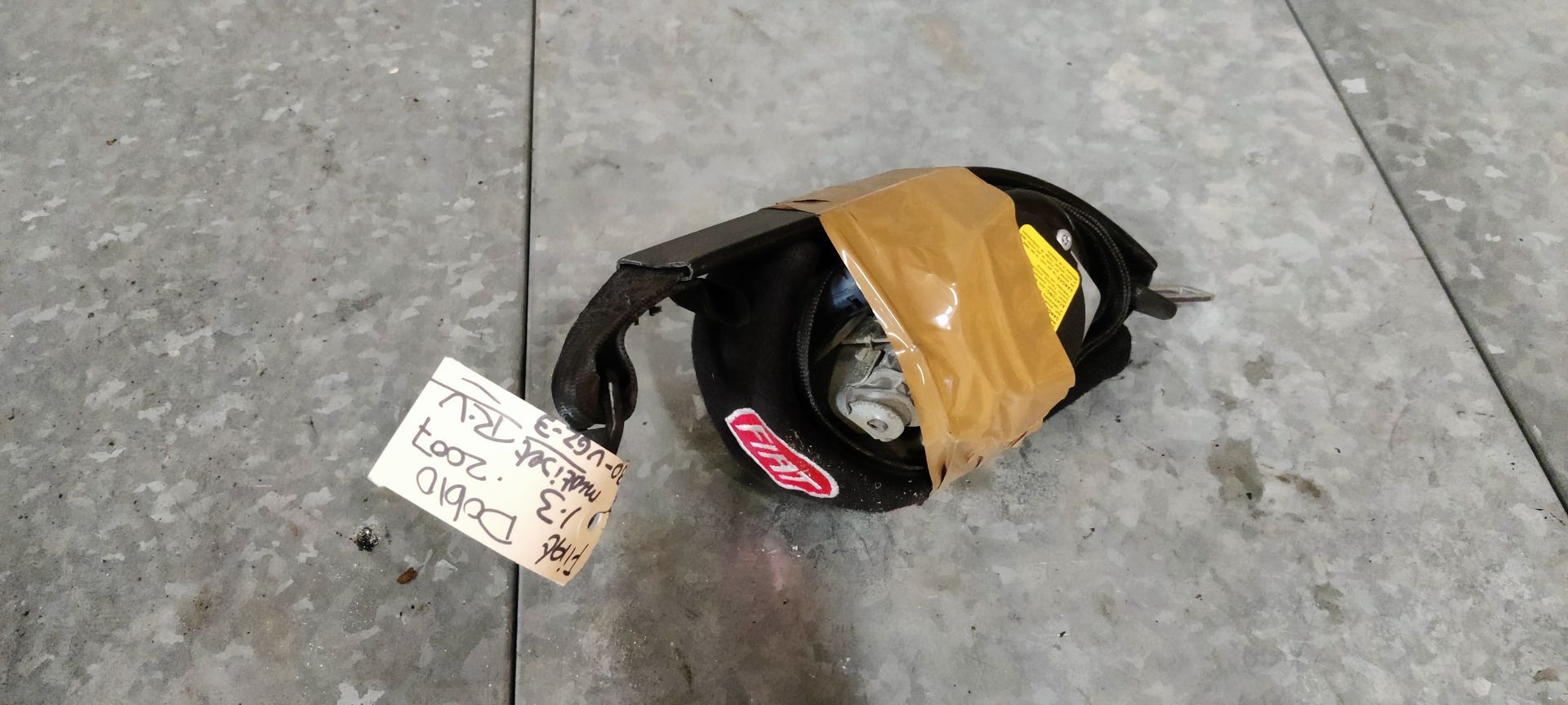 Een oprolmechanisme voor een autogordel, omwikkeld met beige tape, ligt op een grijze vloer met een identificatielabel eraan.