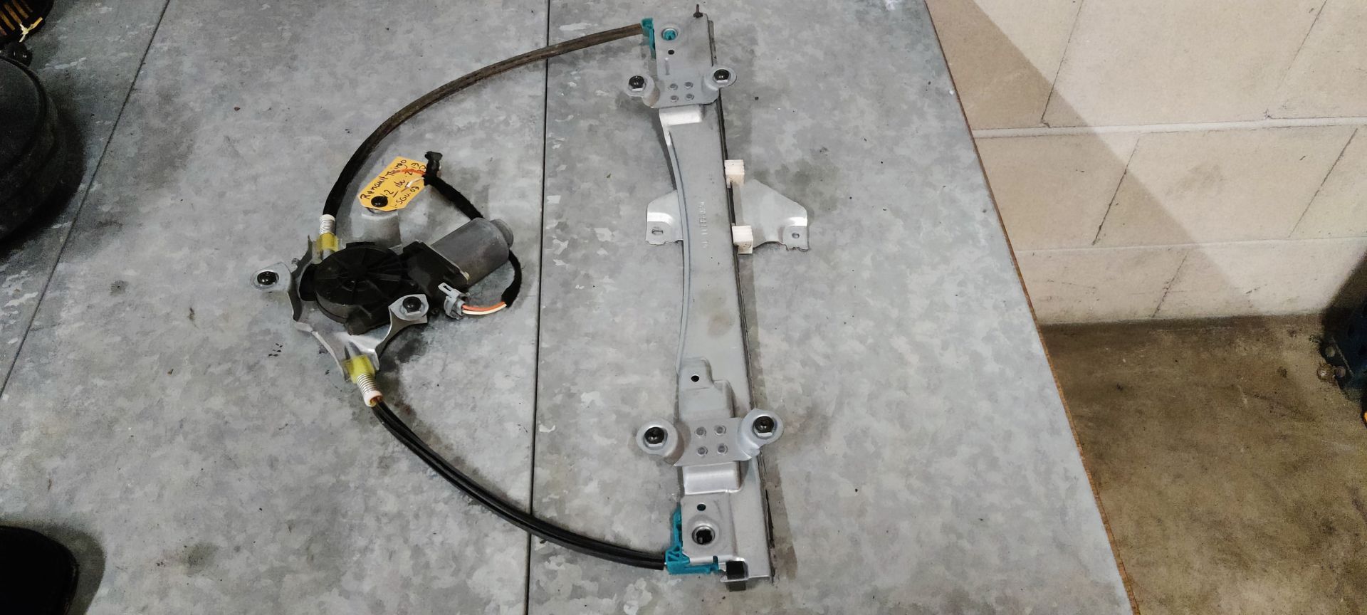 Een zilverkleurige raammechanisme voor auto's, met een zwarte elektromotor en kabel, ligt op een grijs werkblad.