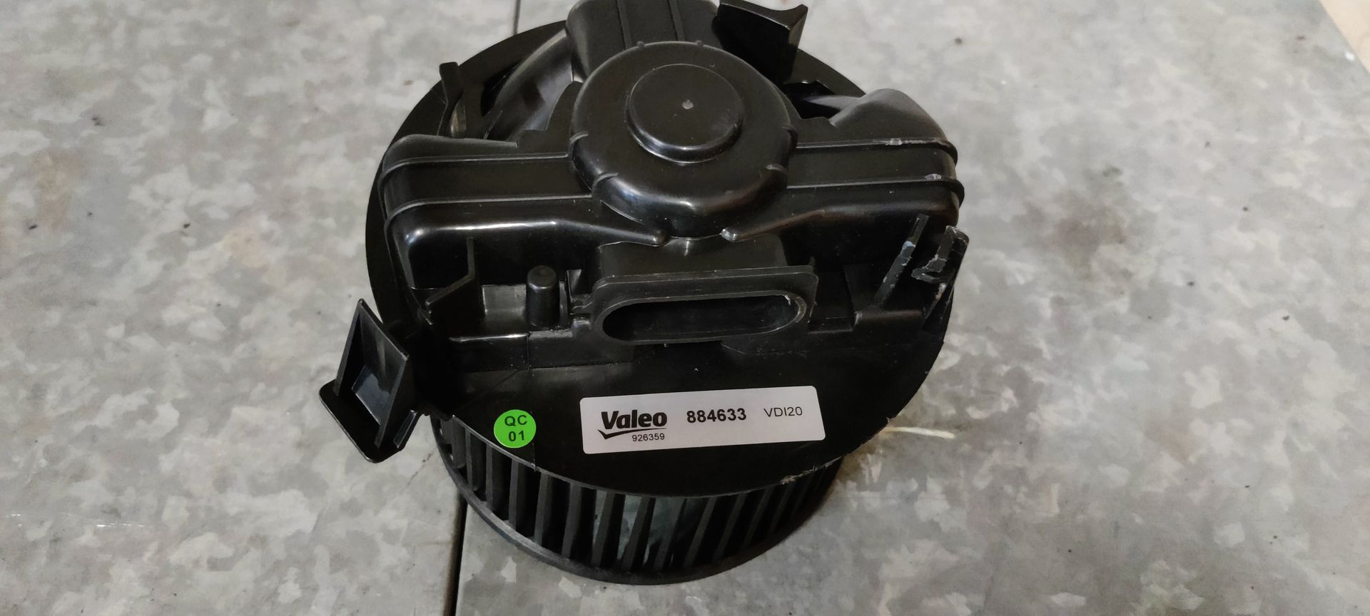 Een zwarte ventilator voor een airconditioningsysteem in een auto, met een ventilatorbehuizing, staat op een grijze vloer.