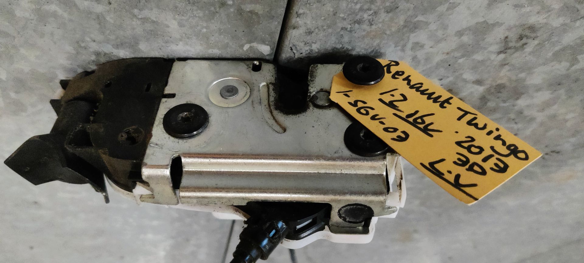 Een metalen deurslotmechanisme voor een Renault Twingo, met een geel identificatielabel op een grijs oppervlak.