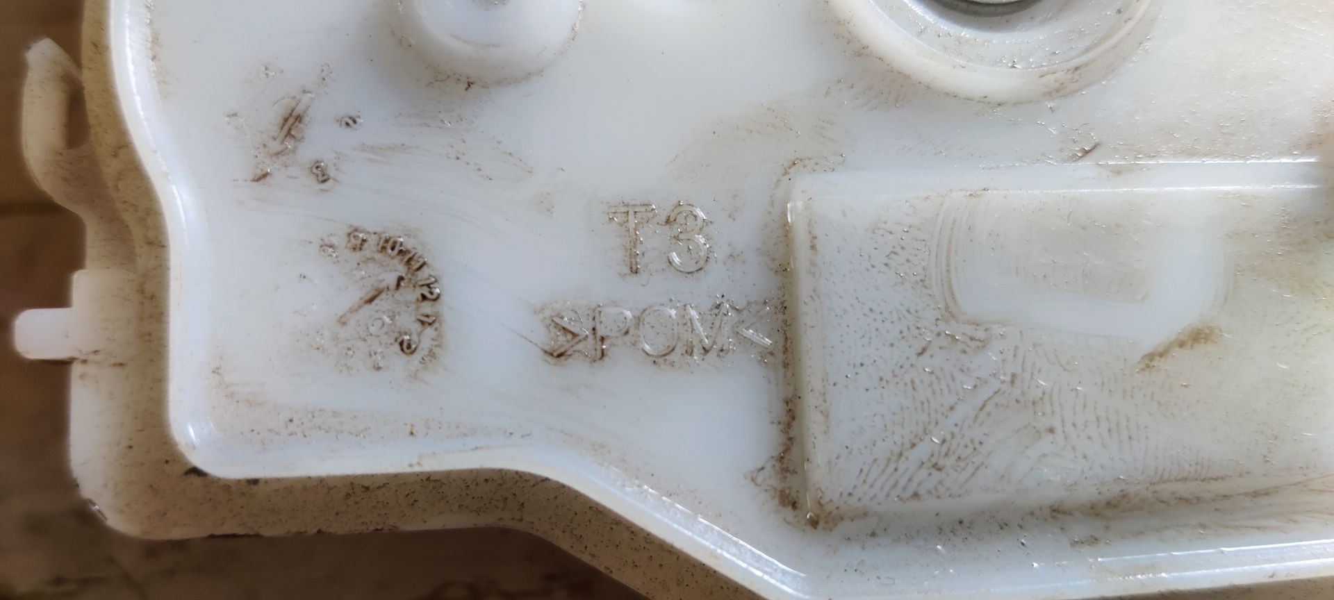 Een zwart metalen kofferbakvergrendelingsmechanisme, bevestigd aan een witte plastic behuizing.
