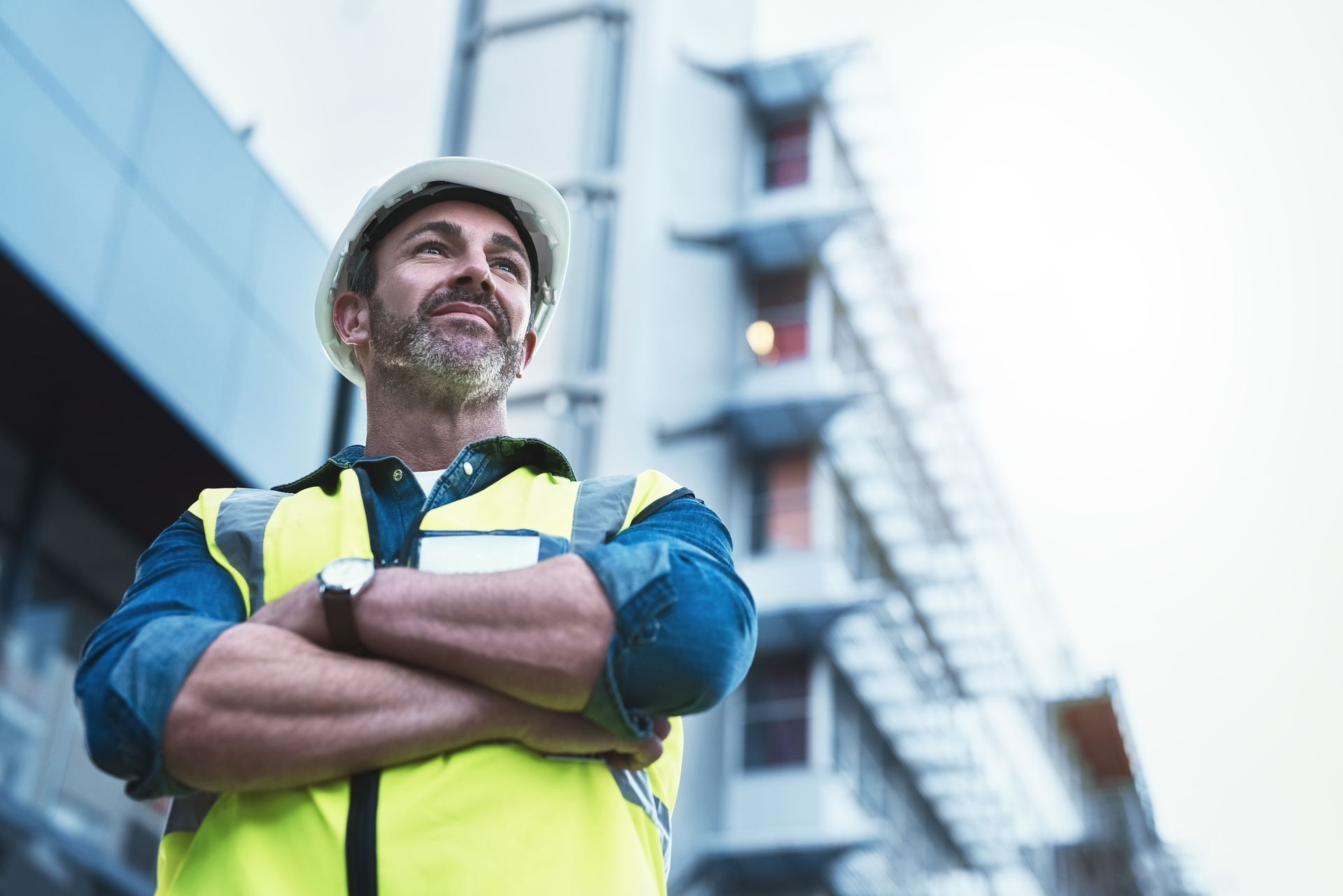 Construction worker in a hard hat and reflective vest standing with arms crossed near a building scaffold