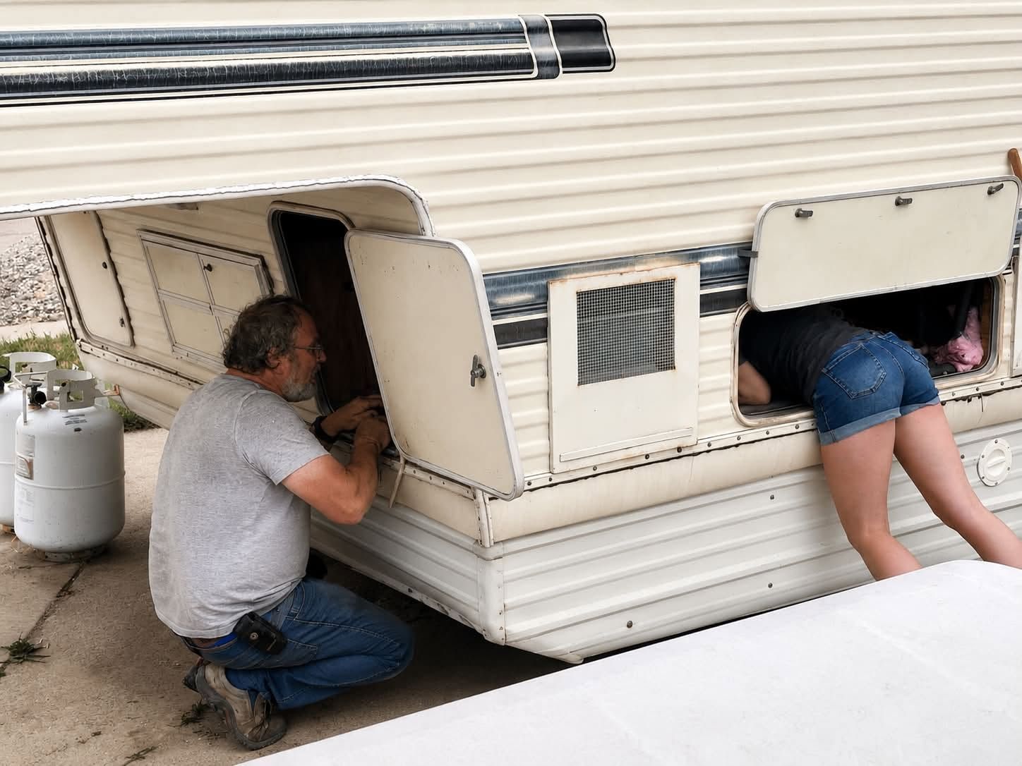 People crawling into a white camper’s storage compartments, one kneeling outside and one legs-first inside