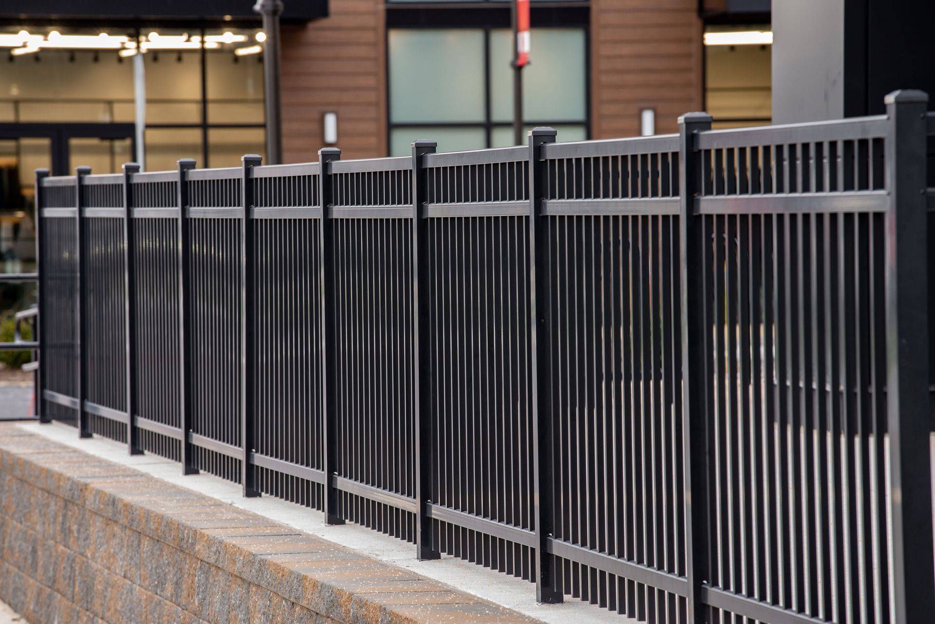 Black metal fence atop a brick wall, in front of a modern building with glass windows Black metal fence atop a brick wall, in front of a modern building with glass windows