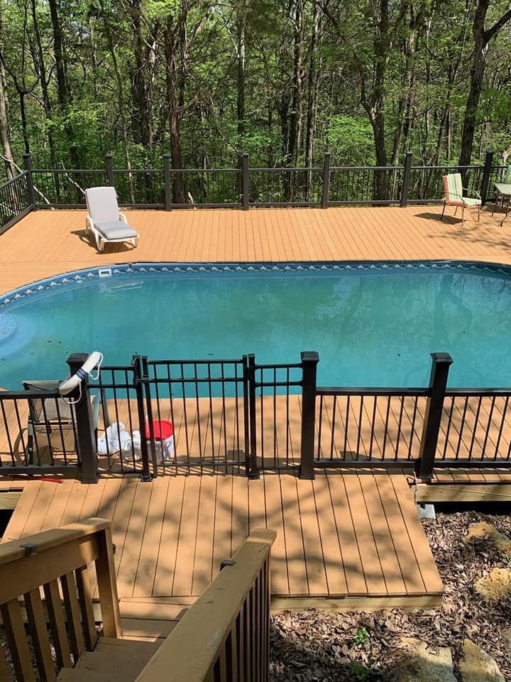 Backyard swimming pool with wooden deck, black fence, and lounge chairs beside wooded trees.