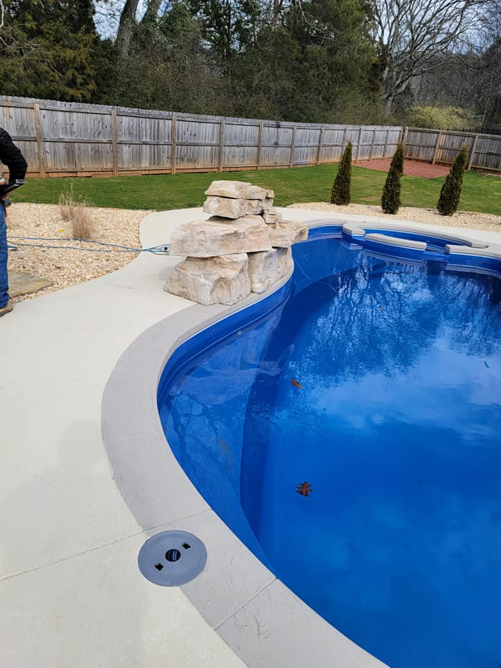 Curved blue backyard pool beside a concrete deck and rock waterfall, with a wooden fence in the background