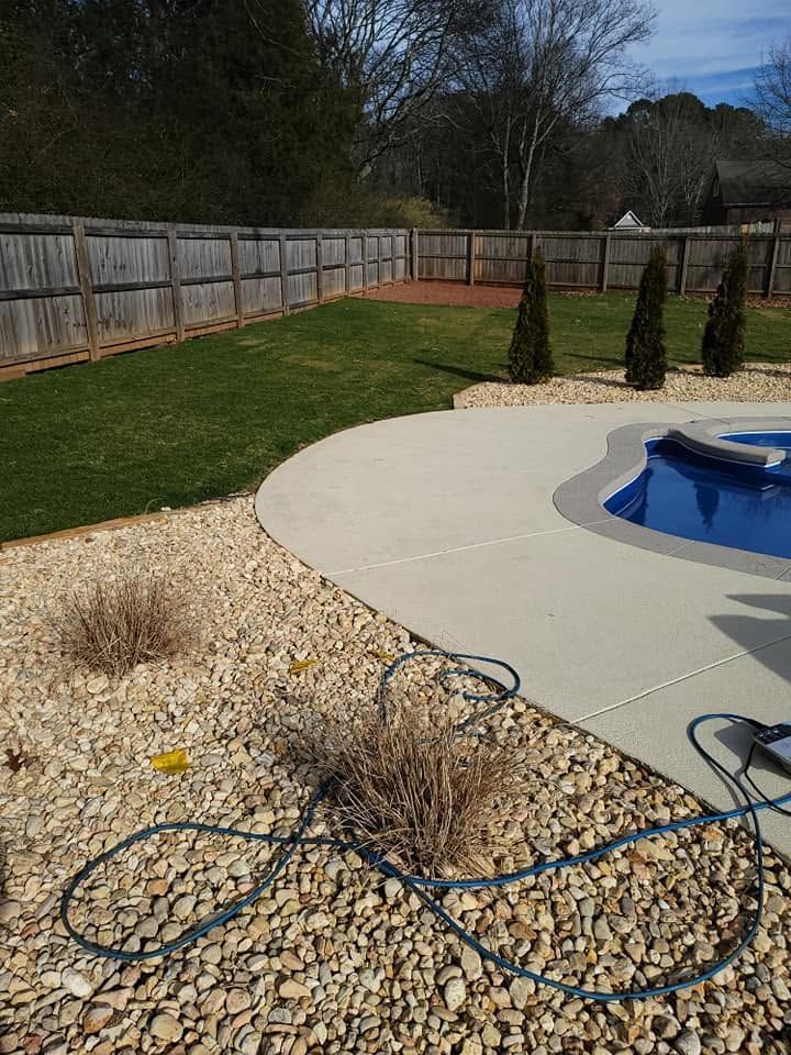 Backyard patio with an in-ground pool, stone edging, grass lawn, fence, and coiled hose in the foreground