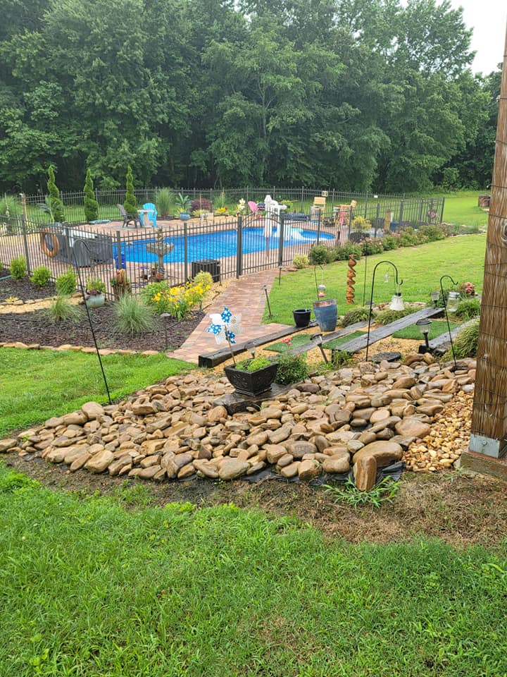 Backyard garden with a blue pool, stone path, and rock waterfall under cloudy skies