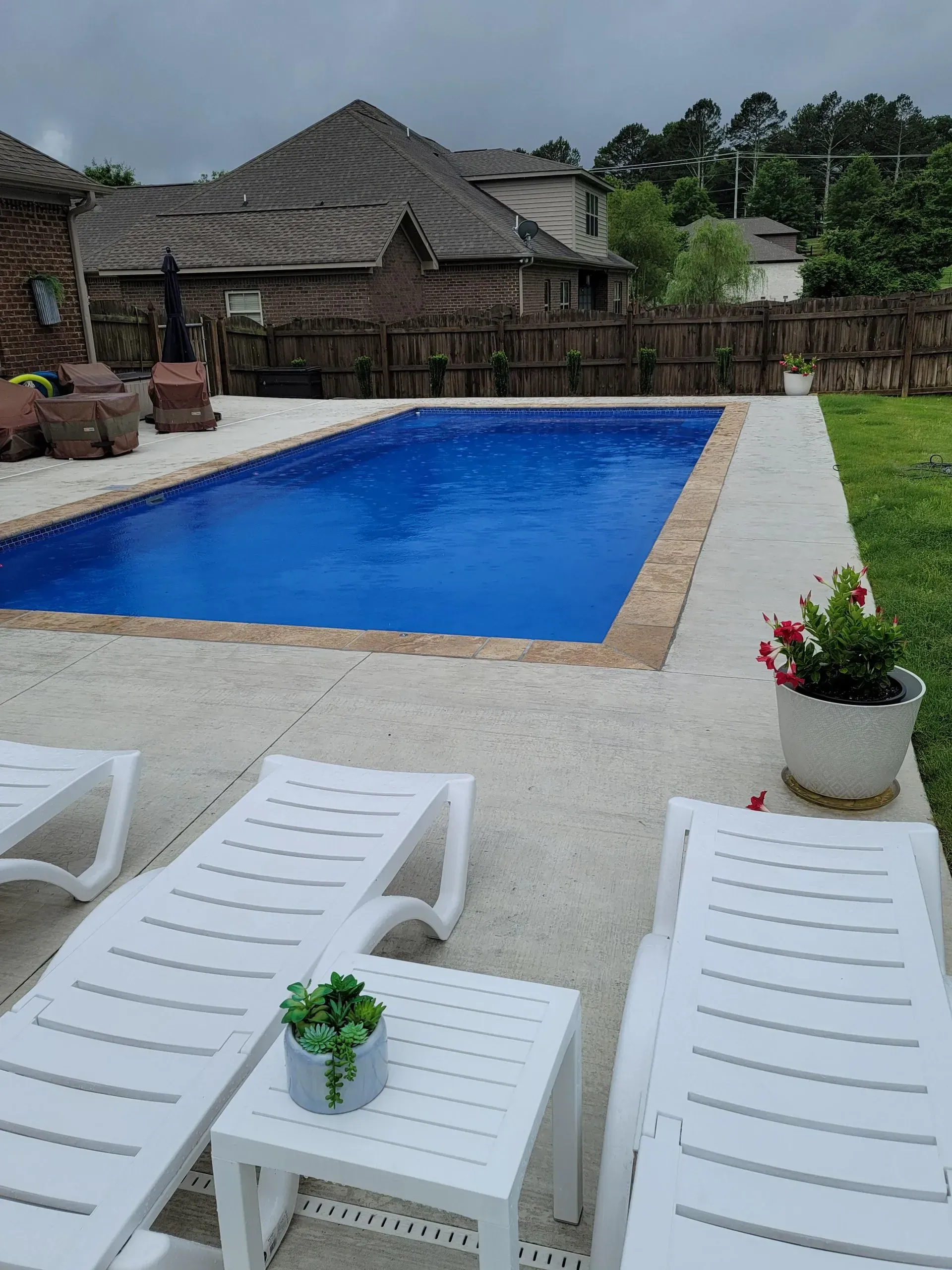 Poolside patio with blue swimming pool, white lounge chairs, and potted plants beside thatched-roof buildings