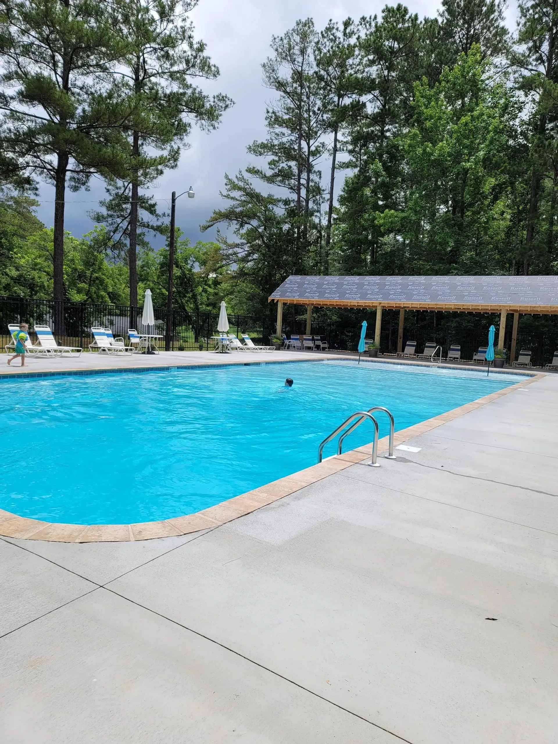 Outdoor swimming pool with blue water, lounge chairs, and trees under a cloudy sky