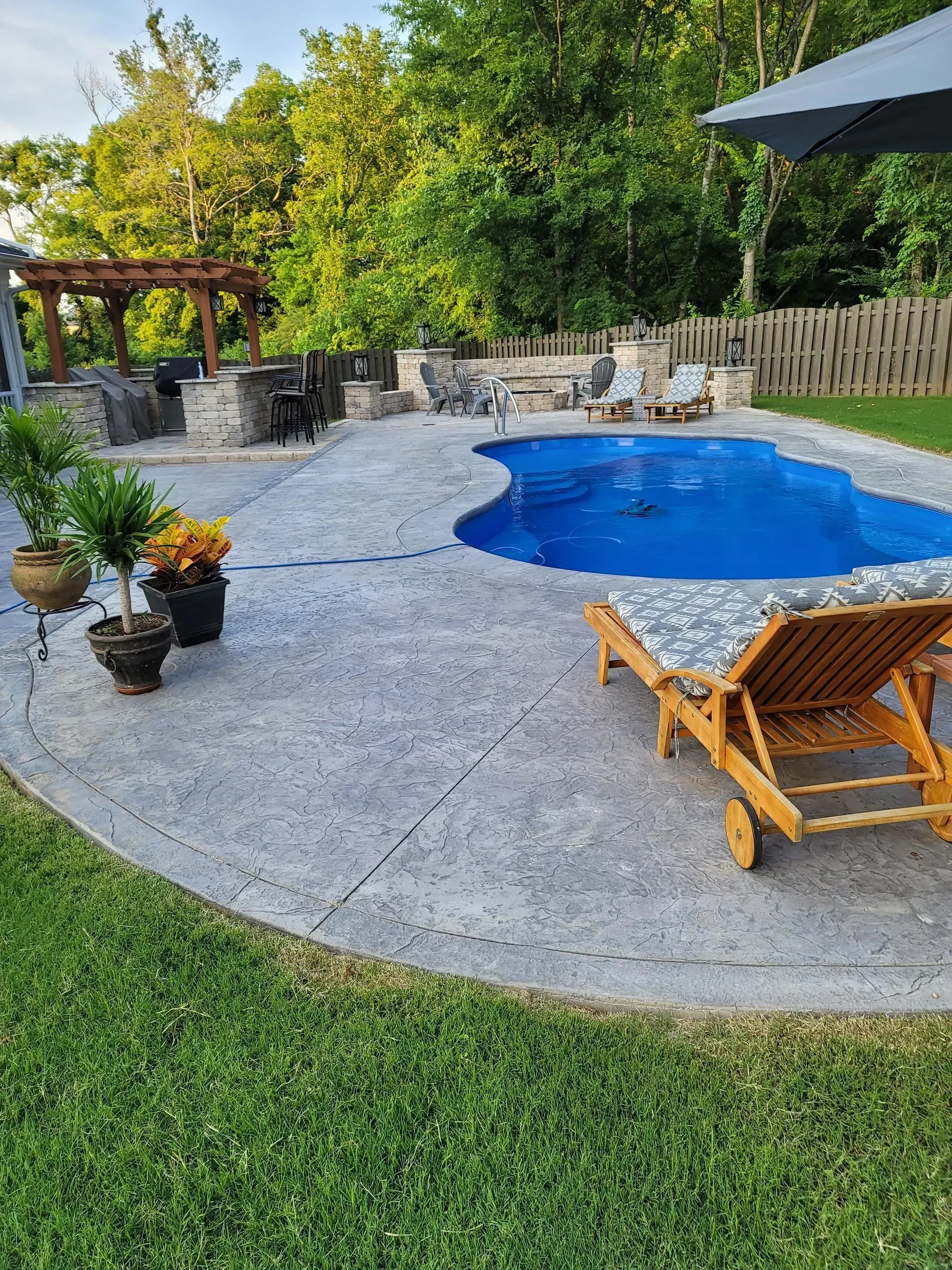 Backyard patio with a blue pool, lounge chair, stone fire pit, and greenery