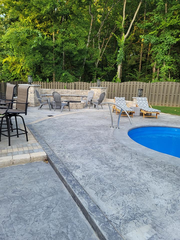 Backyard pool deck with lounge chairs, tables, and a fence beside trees