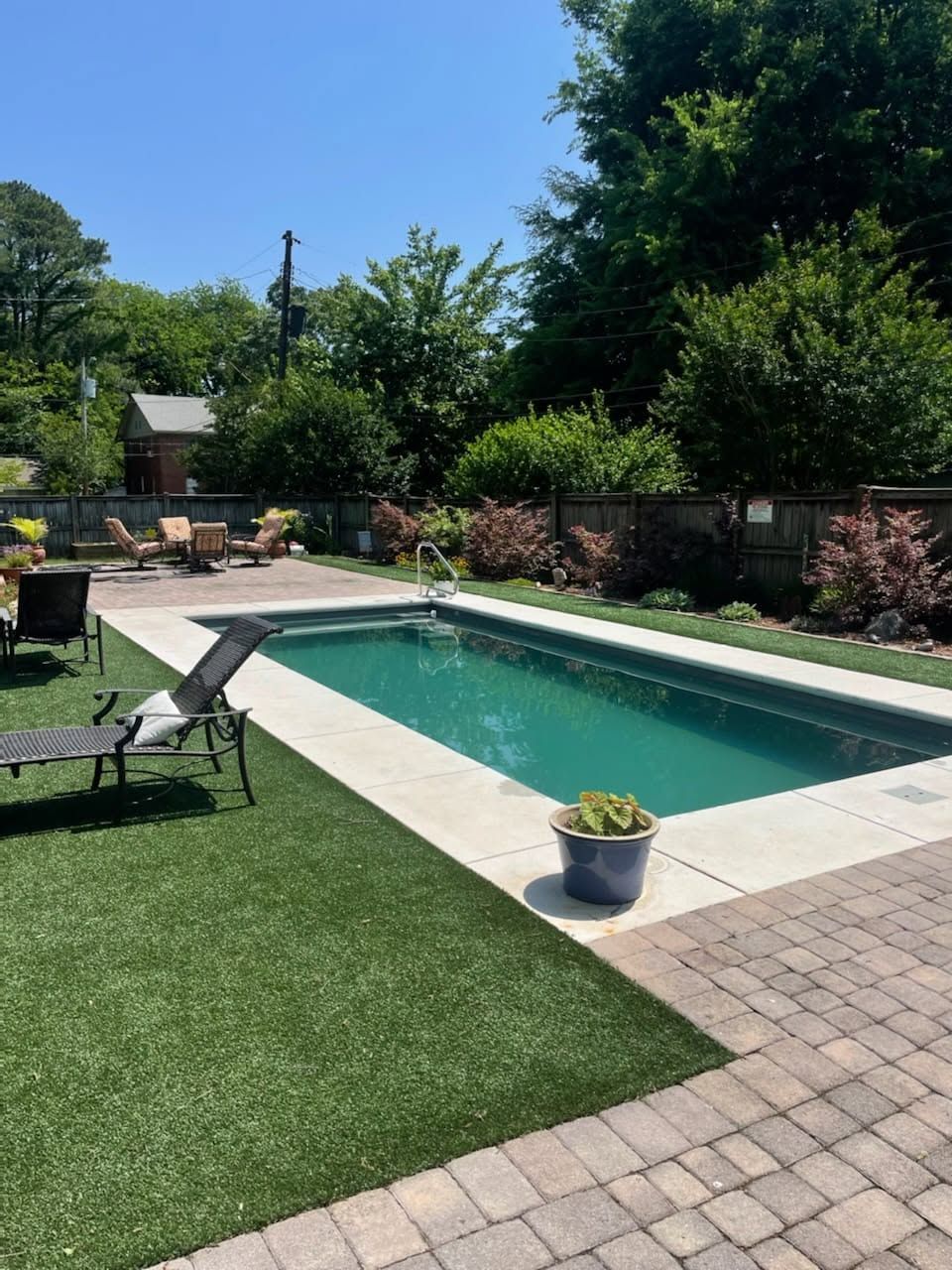 Backyard patio with rectangular swimming pool, lounge chair, and potted plant beside artificial grass.