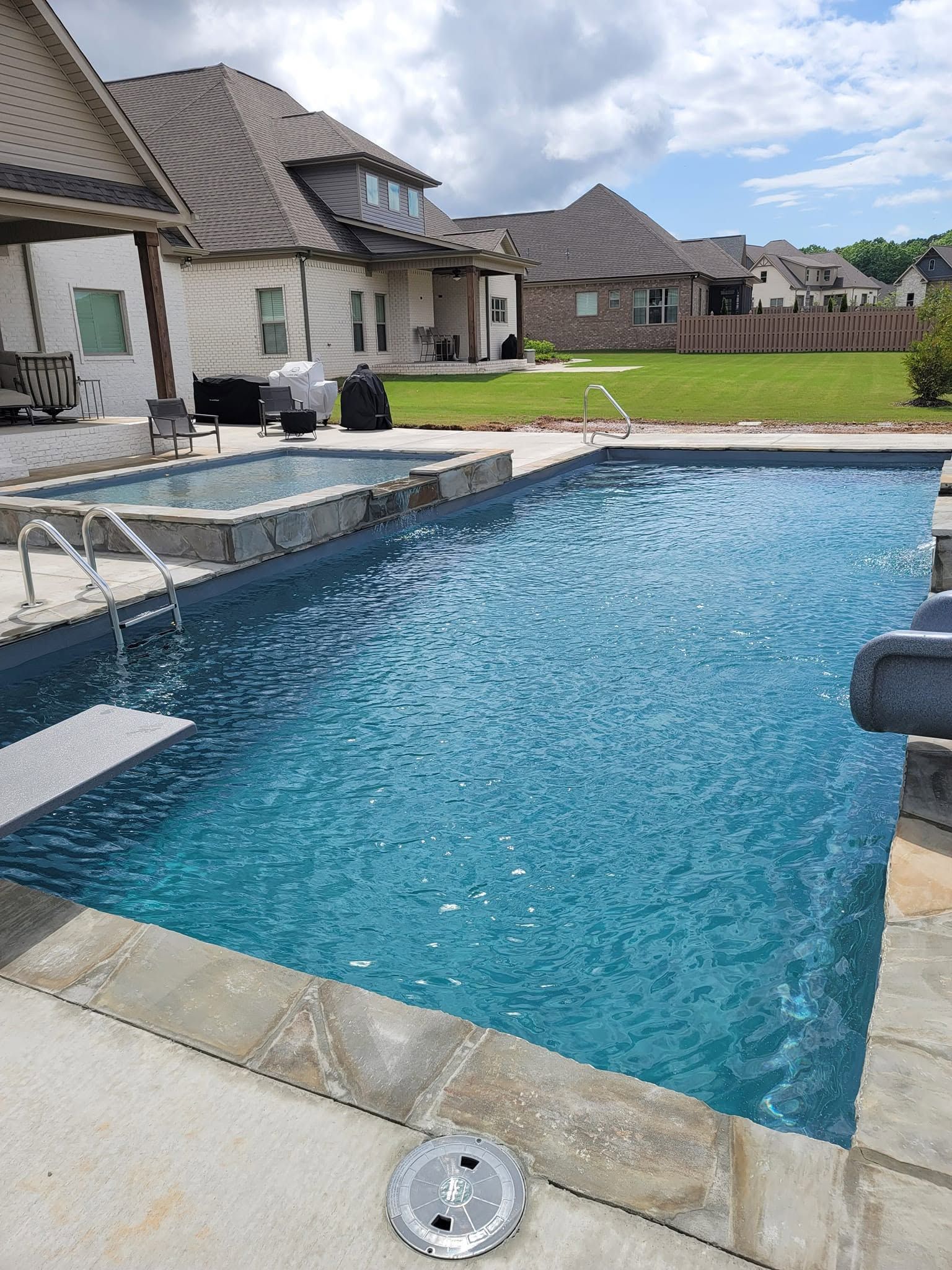 Backyard swimming pool beside a stone patio and suburban houses on a cloudy day