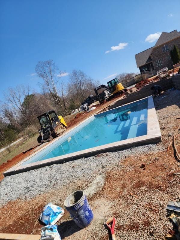 In-ground pool under construction, surrounded by dirt with a small excavator nearby and a house in the background
