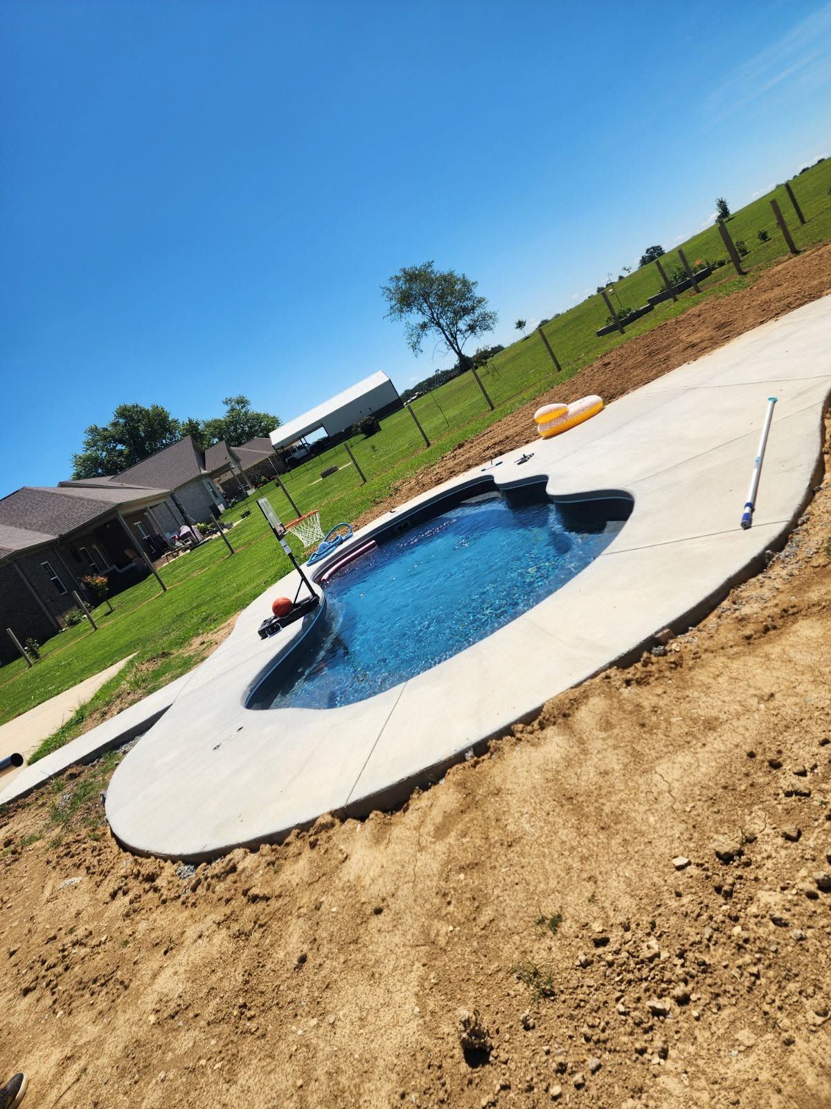 Backyard with a small blue in-ground pool under construction, surrounded by dirt and a white concrete deck