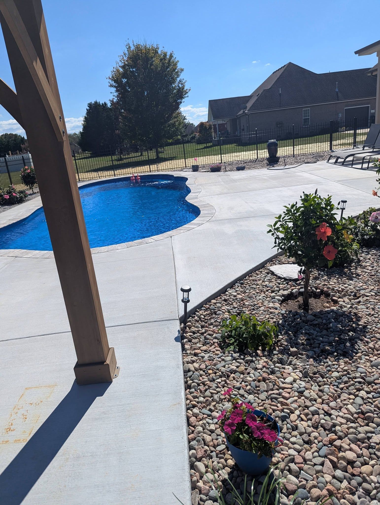 Backyard patio with blue pool, white deck, potted flowers, and a house in bright sun