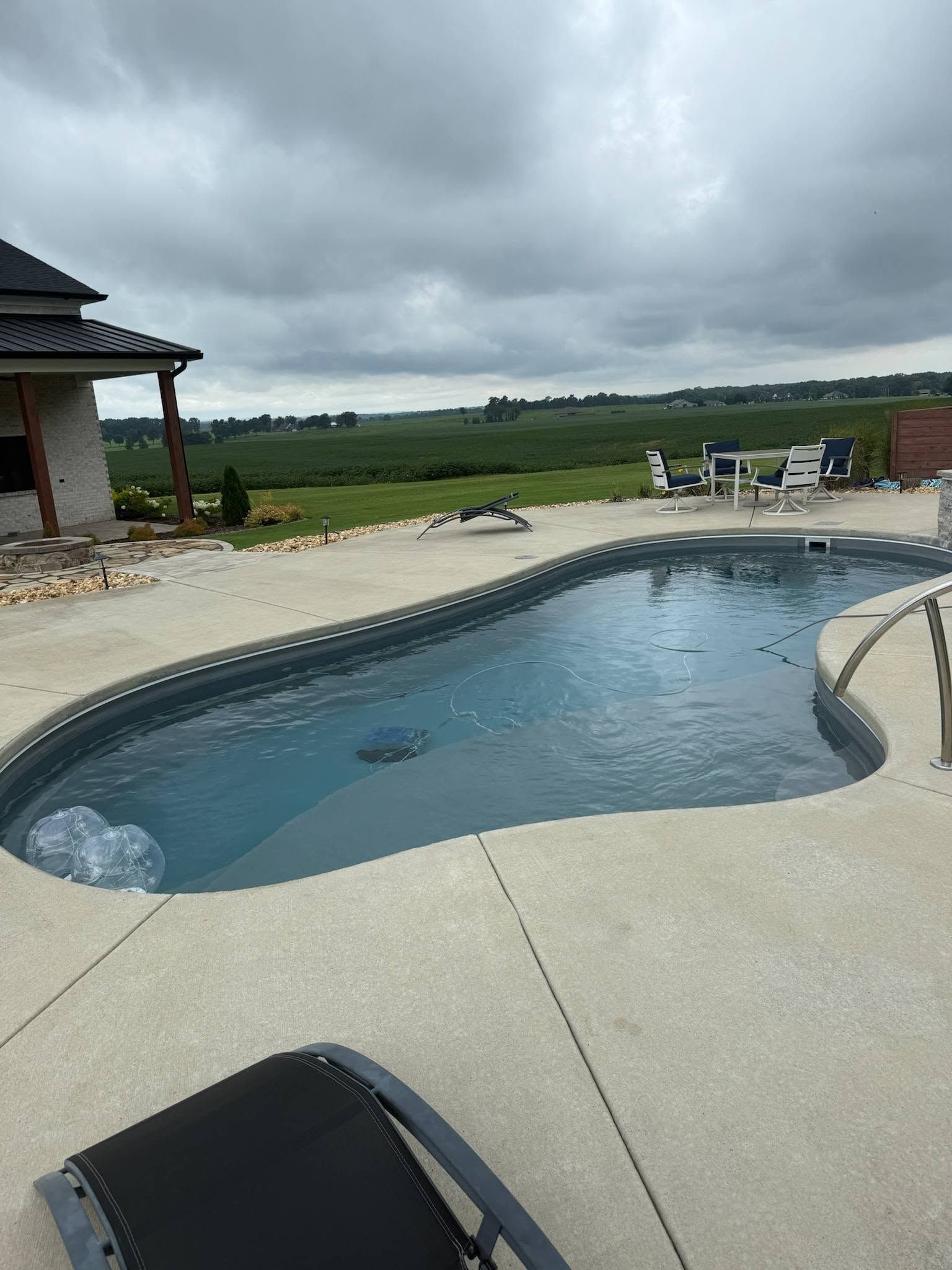 Backyard swimming pool beside a patio with lounge chairs under a cloudy sky.