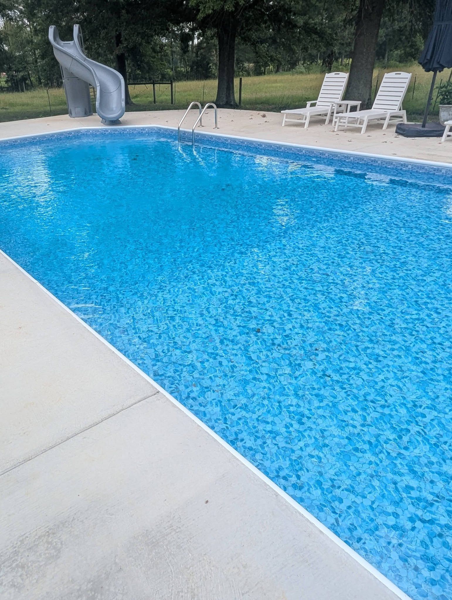 Backyard swimming pool with a gray slide, white lounge chairs, and trees in the background