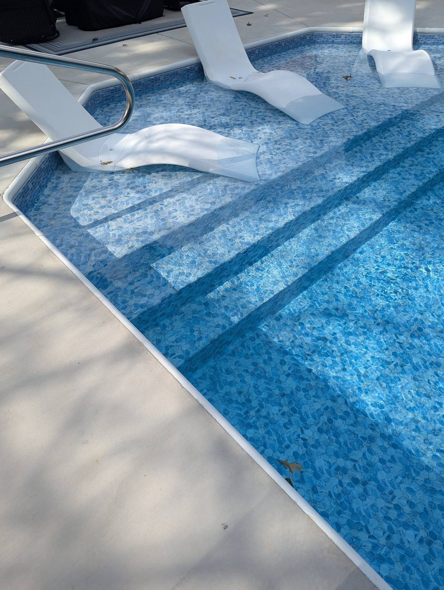 Blue-tiled swimming pool with white poolside lounge chairs and shadows on the deck