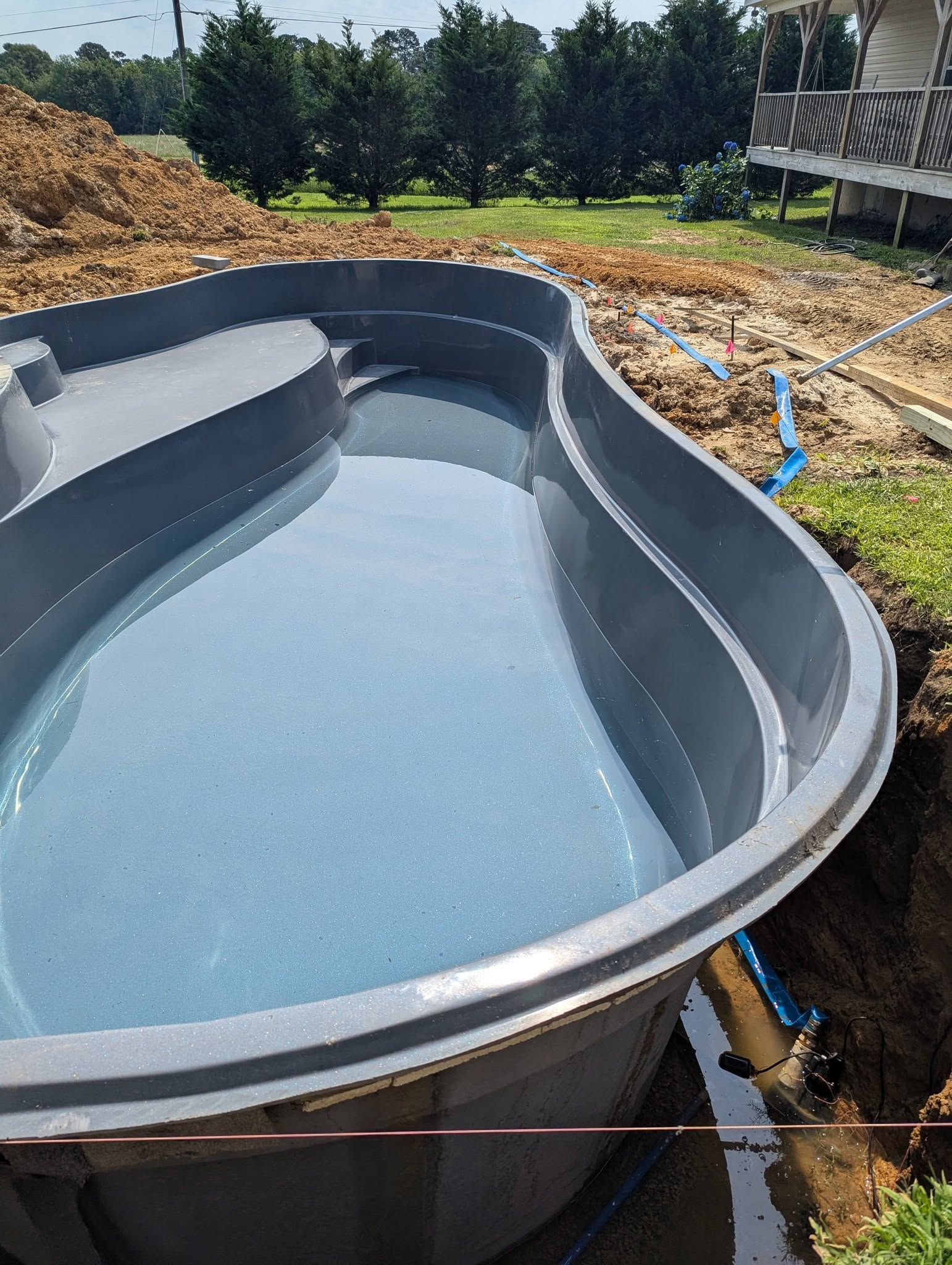 Empty blue pool shell in a backyard construction site, surrounded by dirt and trees