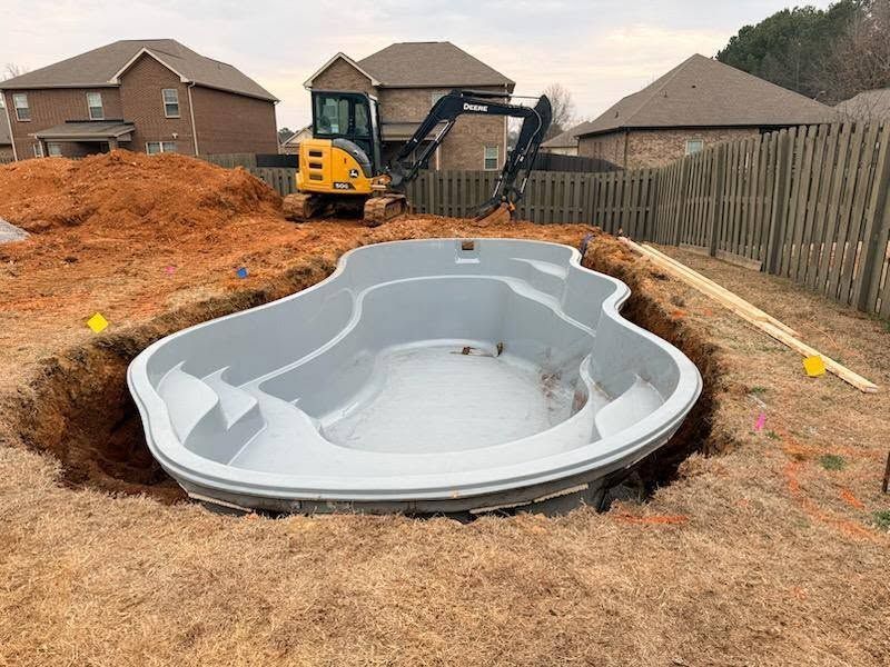 Excavator installing a fiberglass backyard pool shell in a suburban yard with dirt piles and houses nearby